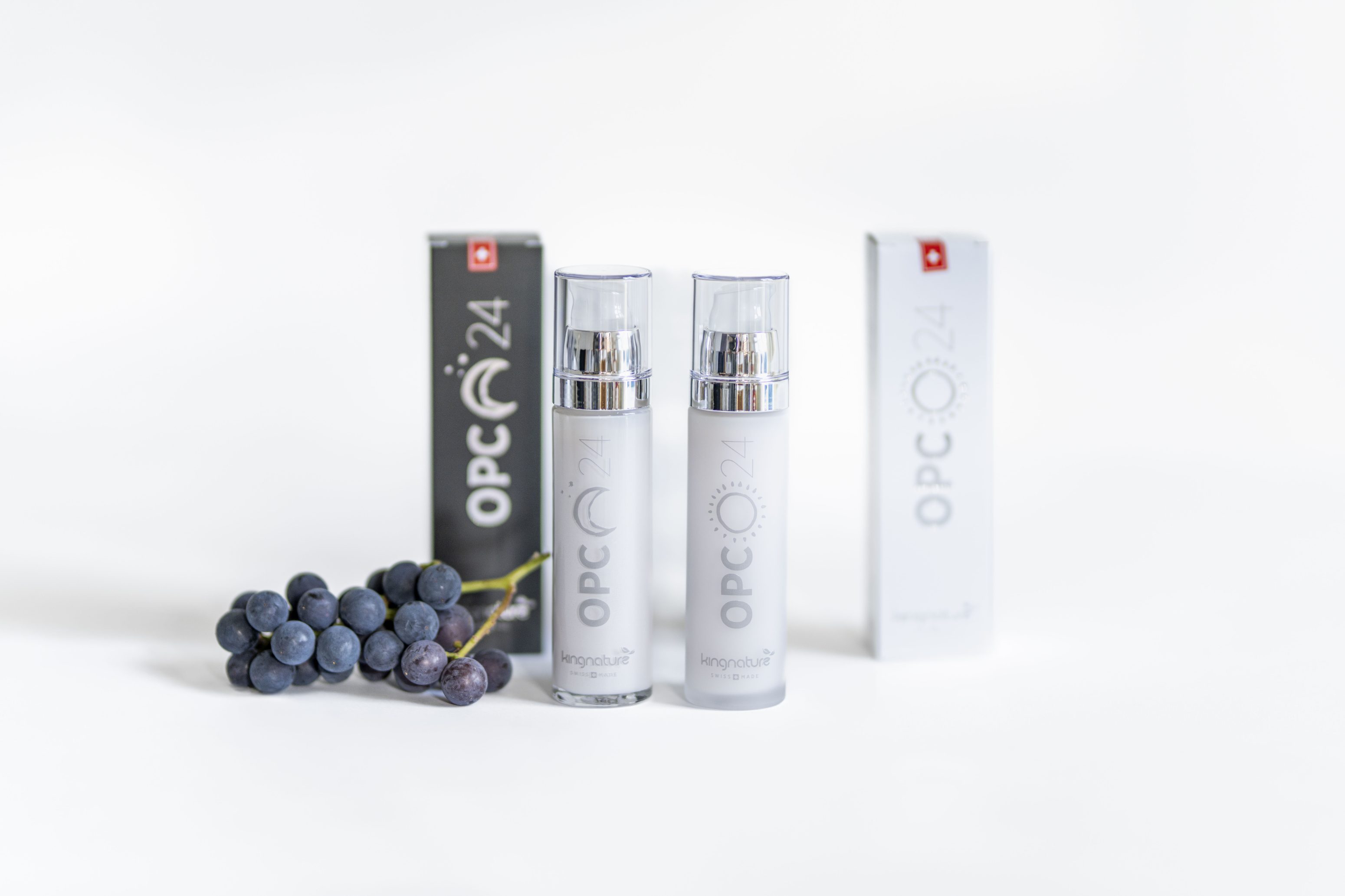 Zwei silberne Flaschen des Hautpflegeprodukts OPC 24 stehen vor ihren Kartons, links davon eine Traube aus dunklen Weintrauben, alles auf weißem Hintergrund.