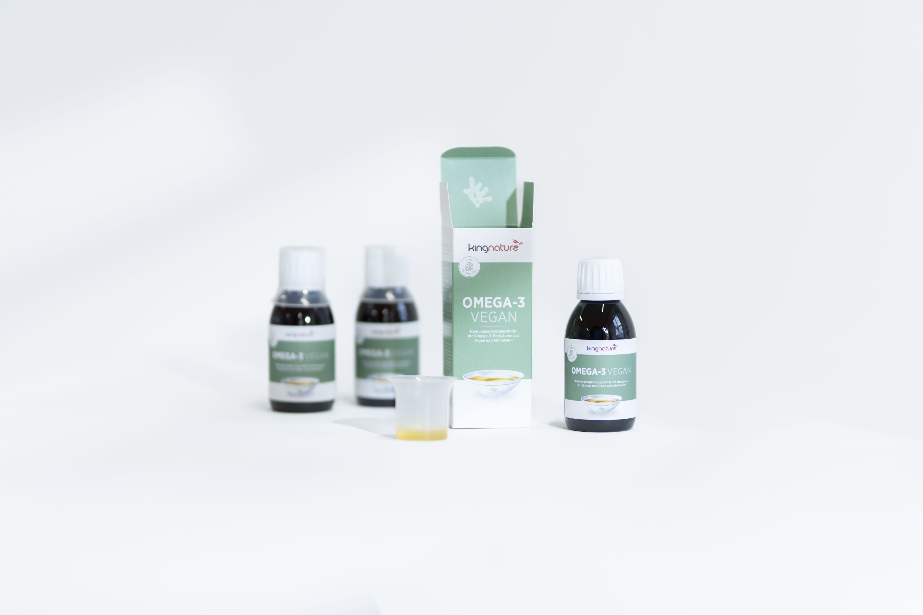 Drei Flaschen und eine Schachtel des veganen Nahrungsergänzungsmittels Kaneka Nutr Omega-3 sind auf einer weißen Fläche ausgestellt, wobei eine geöffnete Flasche, ein Messbecher mit gelber Flüssigkeit und der Verpackungstext sichtbar sind.