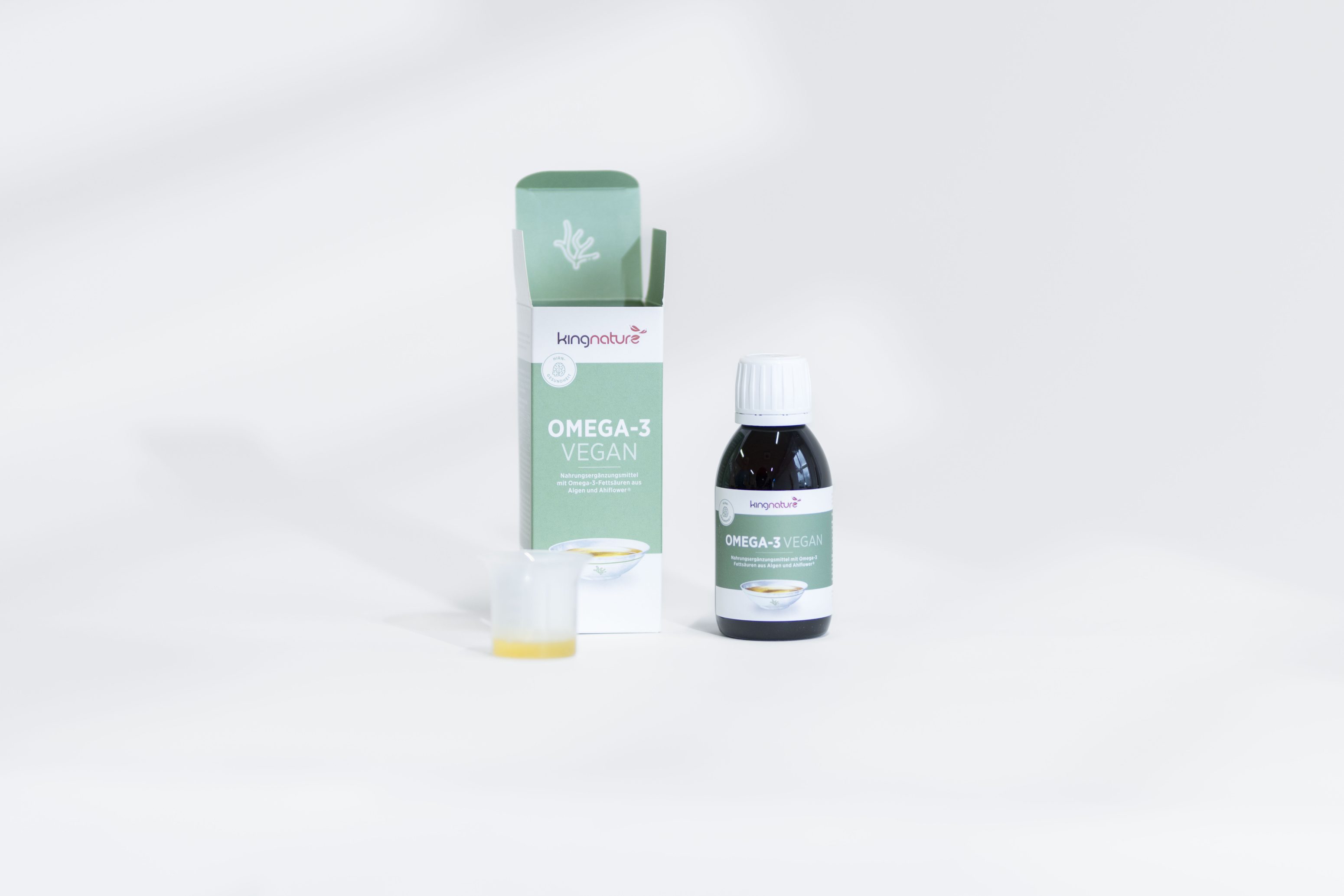 Eine Flasche eines veganen Omega-3-Ergänzungsmittels mit grünem und weißem Etikett steht neben der Verpackungsschachtel und einem kleinen durchsichtigen Messbecher mit gelber Flüssigkeit, alles auf weißem Hintergrund.