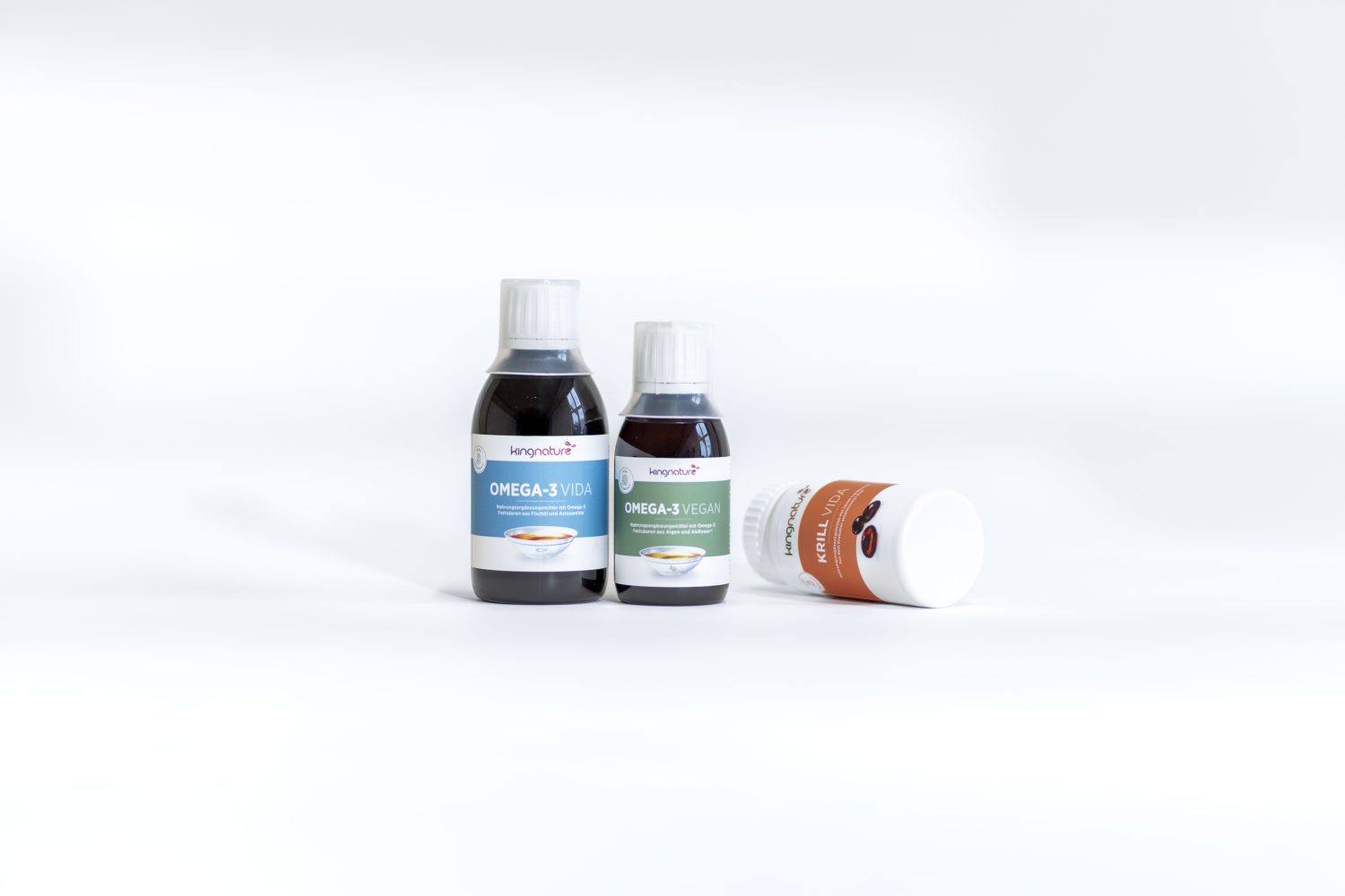 Drei Flaschen mit Nahrungsergänzungsmitteln auf weißem Hintergrund: zwei aufrecht stehende Flaschen mit der Aufschrift "Omega-3 Vegan" und eine orange-weiße Flasche, die auf der Seite liegt und deren Etikett teilweise sichtbar ist.