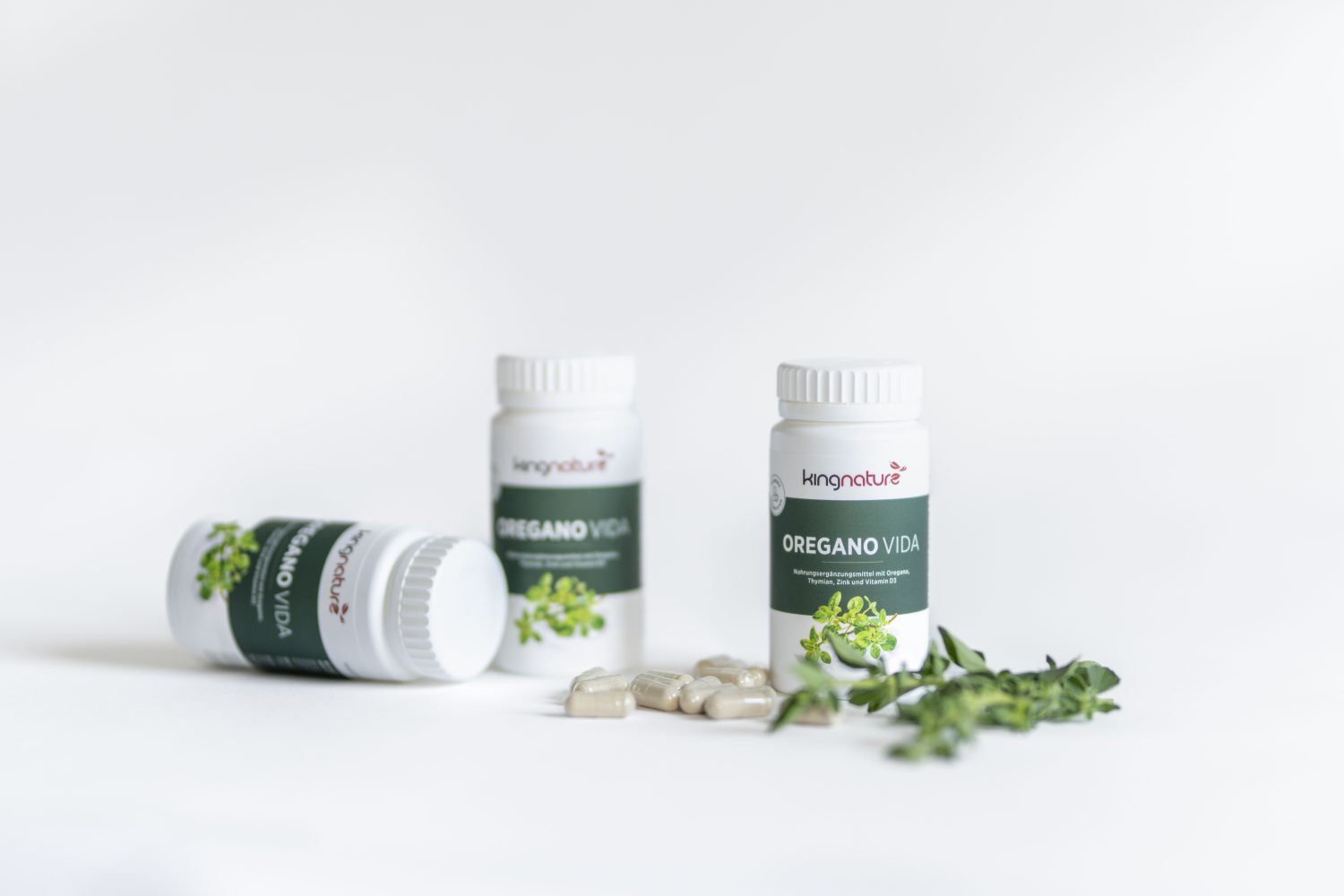 Drei Flaschen Kingnature Oregano Vida Nahrungsergänzungsmittelkapseln, eine liegend und zwei aufrecht stehend, mit losen Kapseln und frischen Oregano-Zweigen, die vor einem weißen Hintergrund verstreut sind.