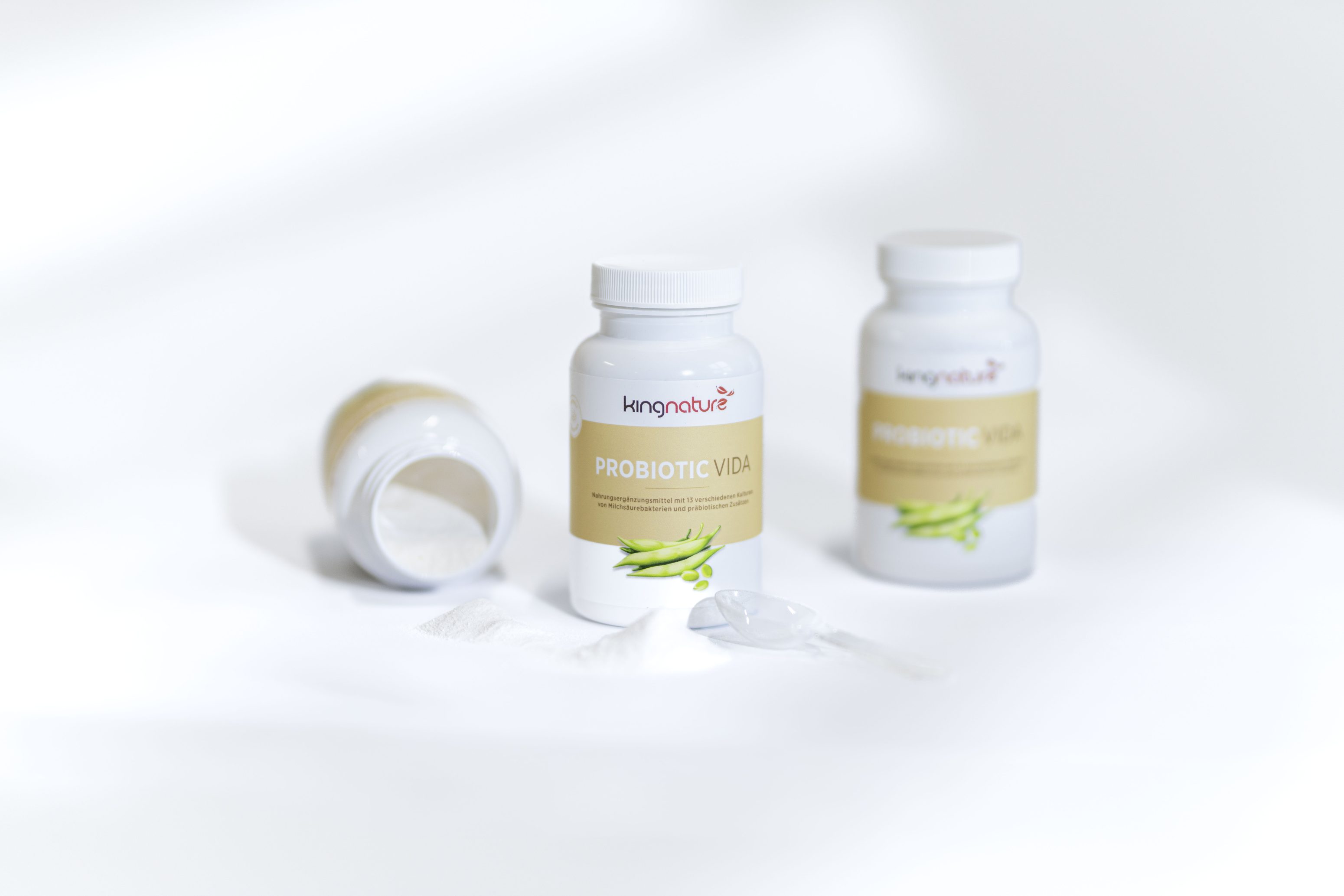 Abgebildet sind drei weiße Flaschen mit Kingnature Probiotic Vida Nahrungsergänzungsmitteln, von denen eine auf der Seite liegt und das Pulver herausläuft, während zwei aufrecht auf einer weißen Fläche stehen. Vorne sind ein durchsichtiger Schöpflöffel und das Pulver zu sehen.