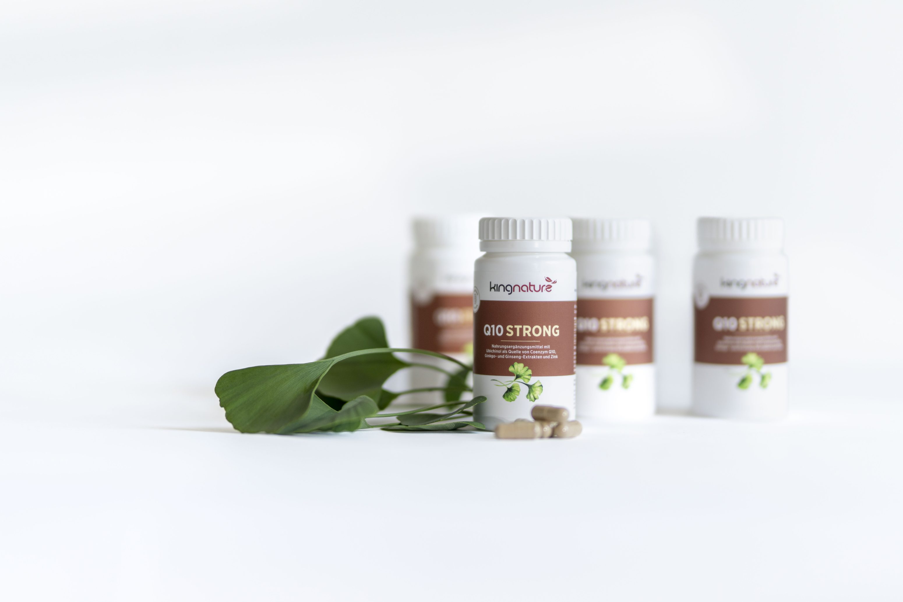 Vier Flaschen des Nahrungsergänzungsmittels Kingnature Q10 Strong sind auf einer weißen Fläche mit grünen Blättern und mehreren beigen Kapseln vor den Flaschen angeordnet.