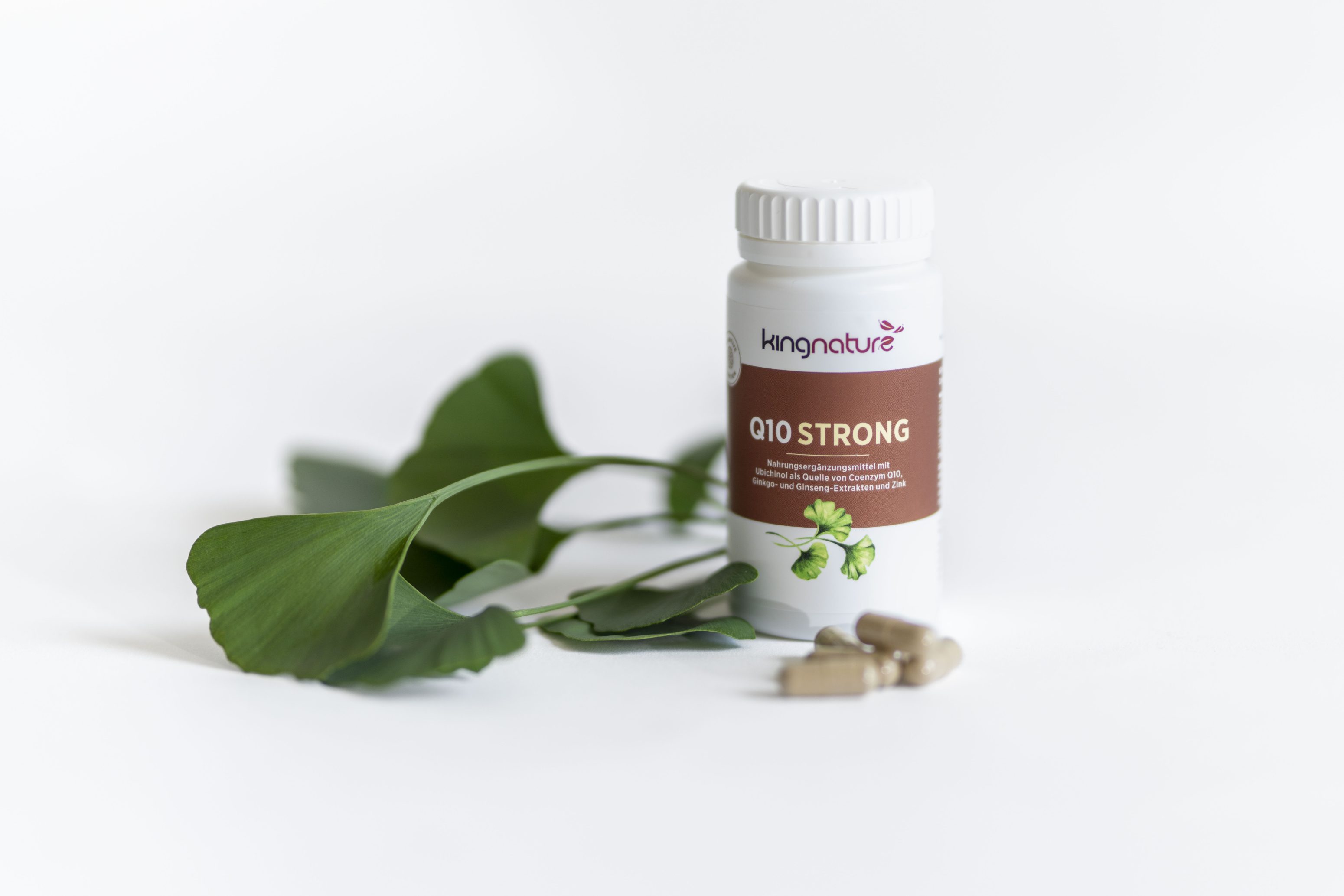 Eine weiße Flasche mit der Aufschrift kingnature Q10 STRONG und einer grünen Blattgrafik steht neben frischen grünen Blättern und drei braunen Kapseln, alles auf weißem Hintergrund.