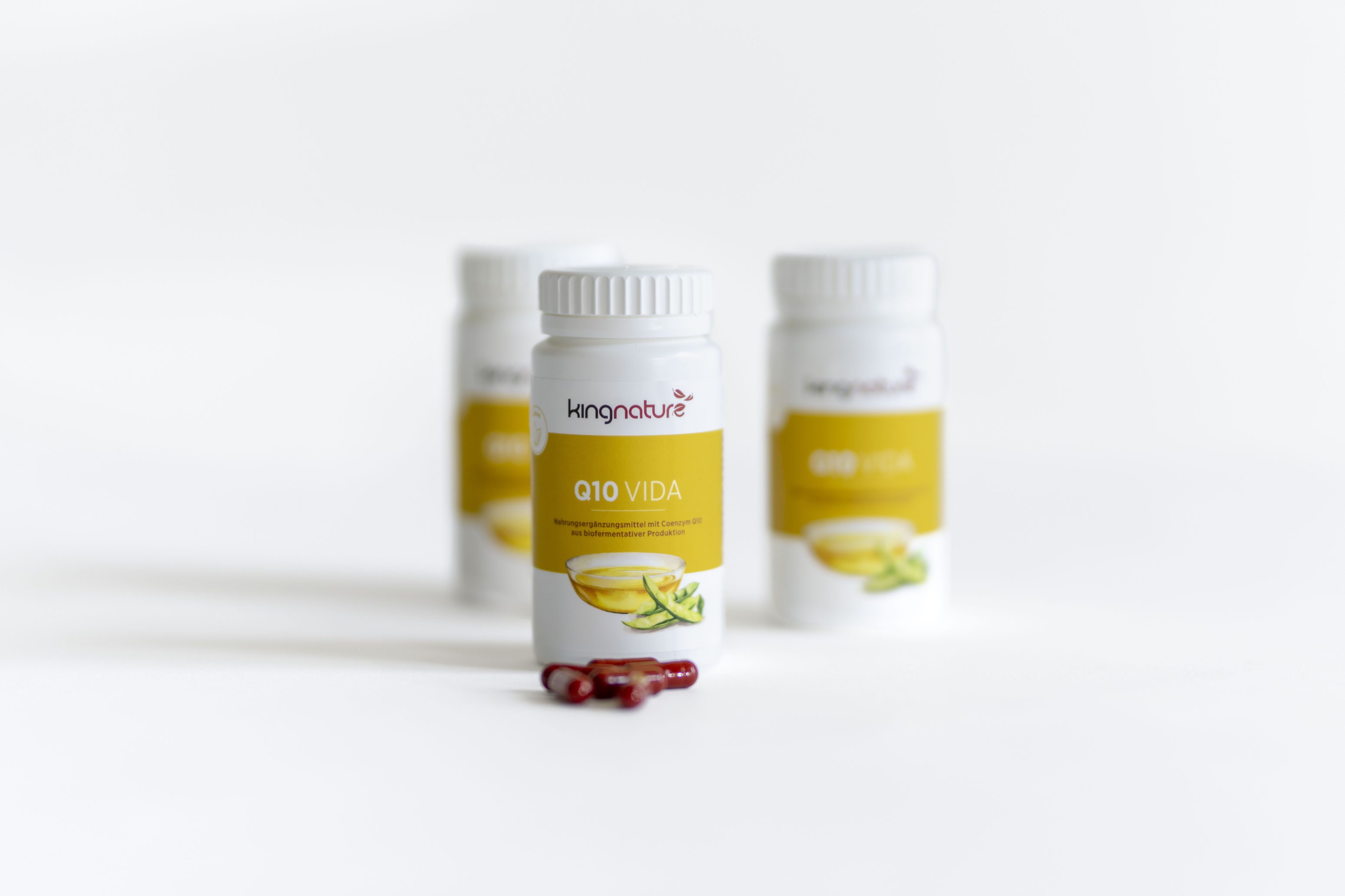 Eine weiße Flasche mit der Aufschrift Q10 VIDA von Kingnature steht im Mittelpunkt, während zwei ähnliche Flaschen im Hintergrund verschwommen sind. Mehrere rote Kapseln liegen vor der Hauptflasche auf einer weißen Fläche.