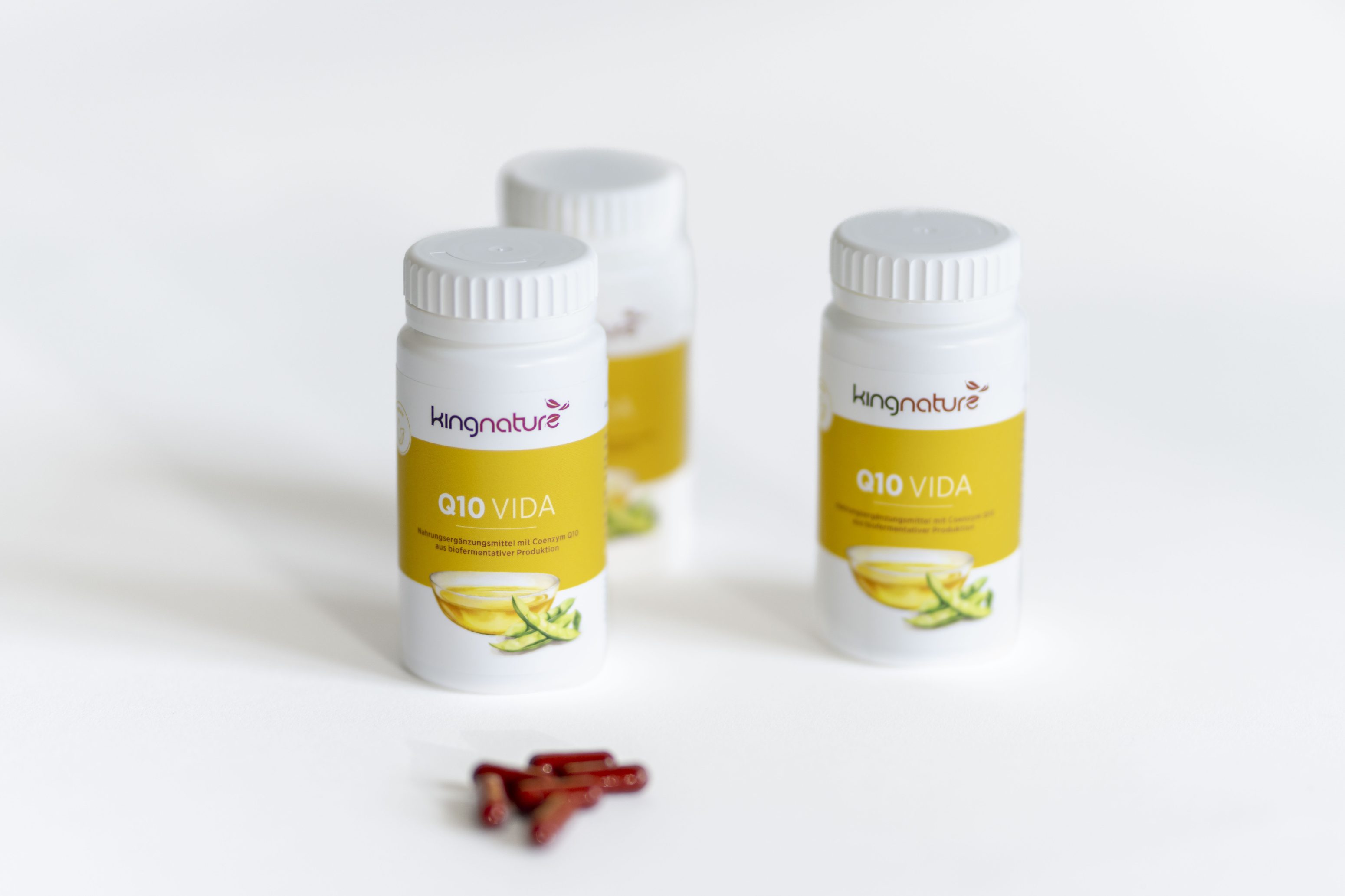 Drei weiße Flaschen mit der Aufschrift kingnature Q10 VIDA und gelben Etiketten stehen auf einer weißen Fläche, davor liegen vier rote Kapseln. Die Flaschen und Kapseln deuten auf ein Nahrungsergänzungsprodukt hin.