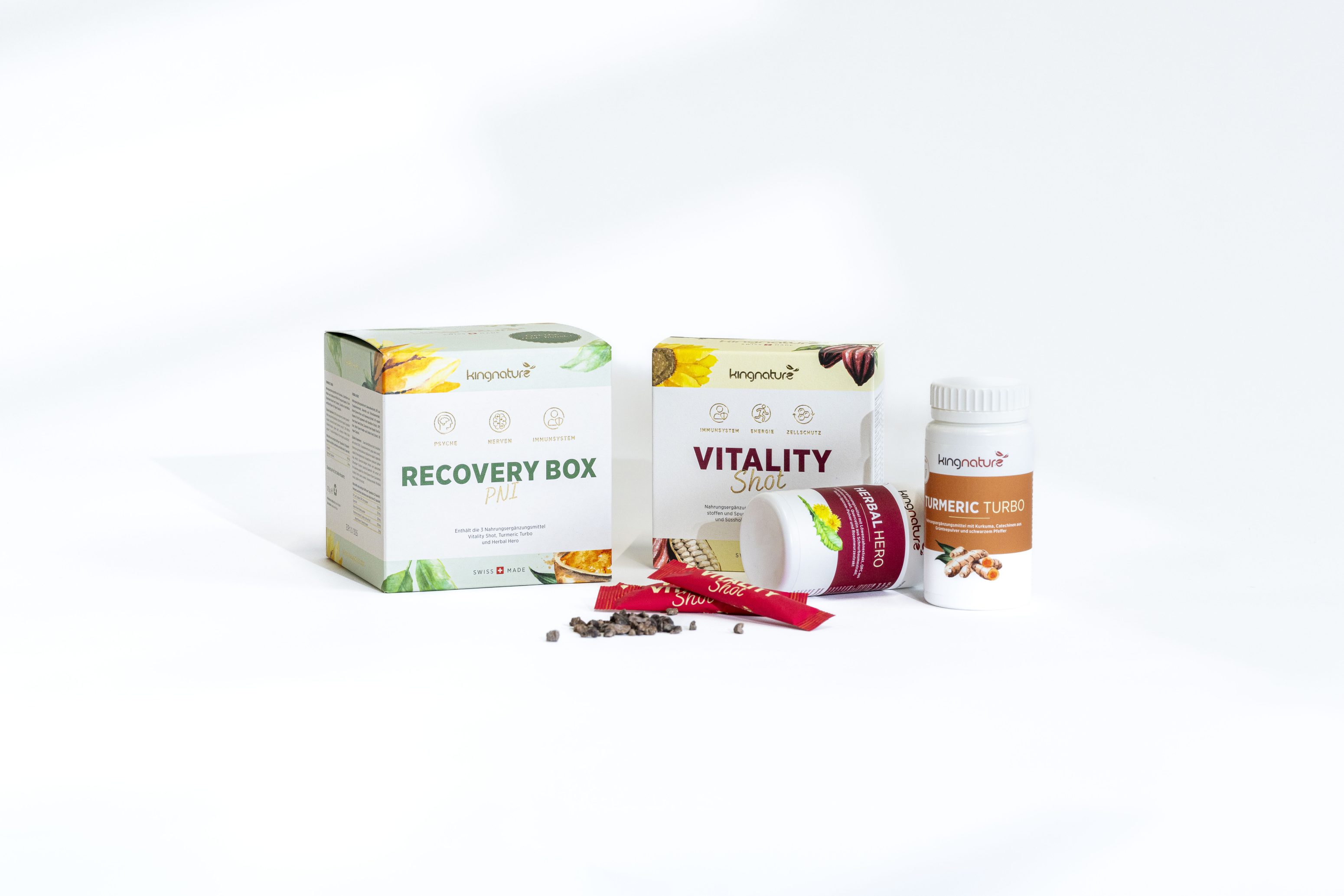 Auf einem weißen Hintergrund sind Schachteln mit den Aufschriften "Recovery Box Pill" und "Vitality Shot", eine weiße Flasche mit der Aufschrift "Curcumin Turmeric", zwei rote Stick-Pakete und lose Pillen angeordnet.