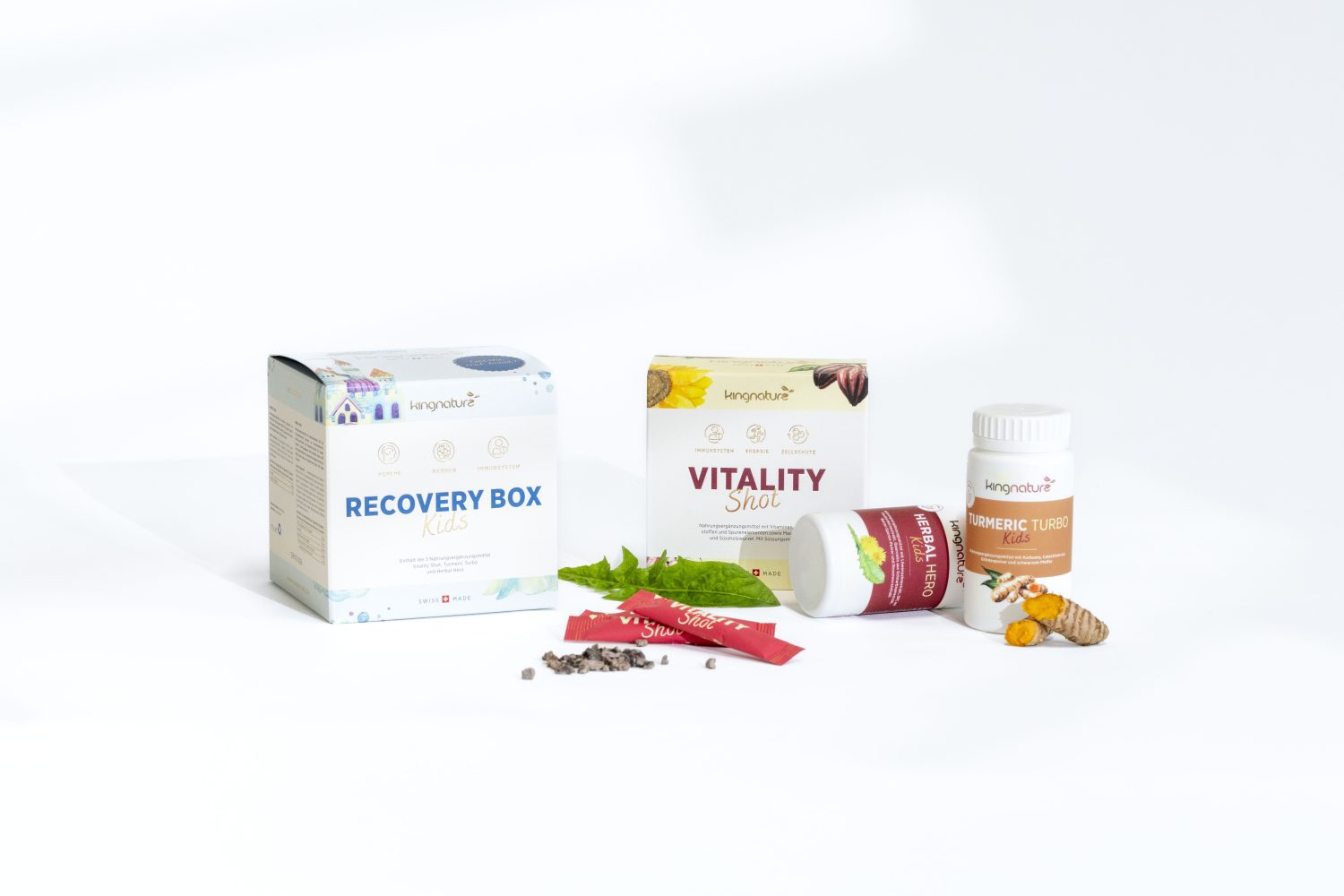 Drei Wellness-Produkte sind ausgestellt: eine Recovery Box Kids, ein Vitality Shot und eine Flasche Kurkuma Tumeric. Frische Zutaten und Ergänzungsstäbchen sind auf weißem Hintergrund um die Produkte herum angeordnet.