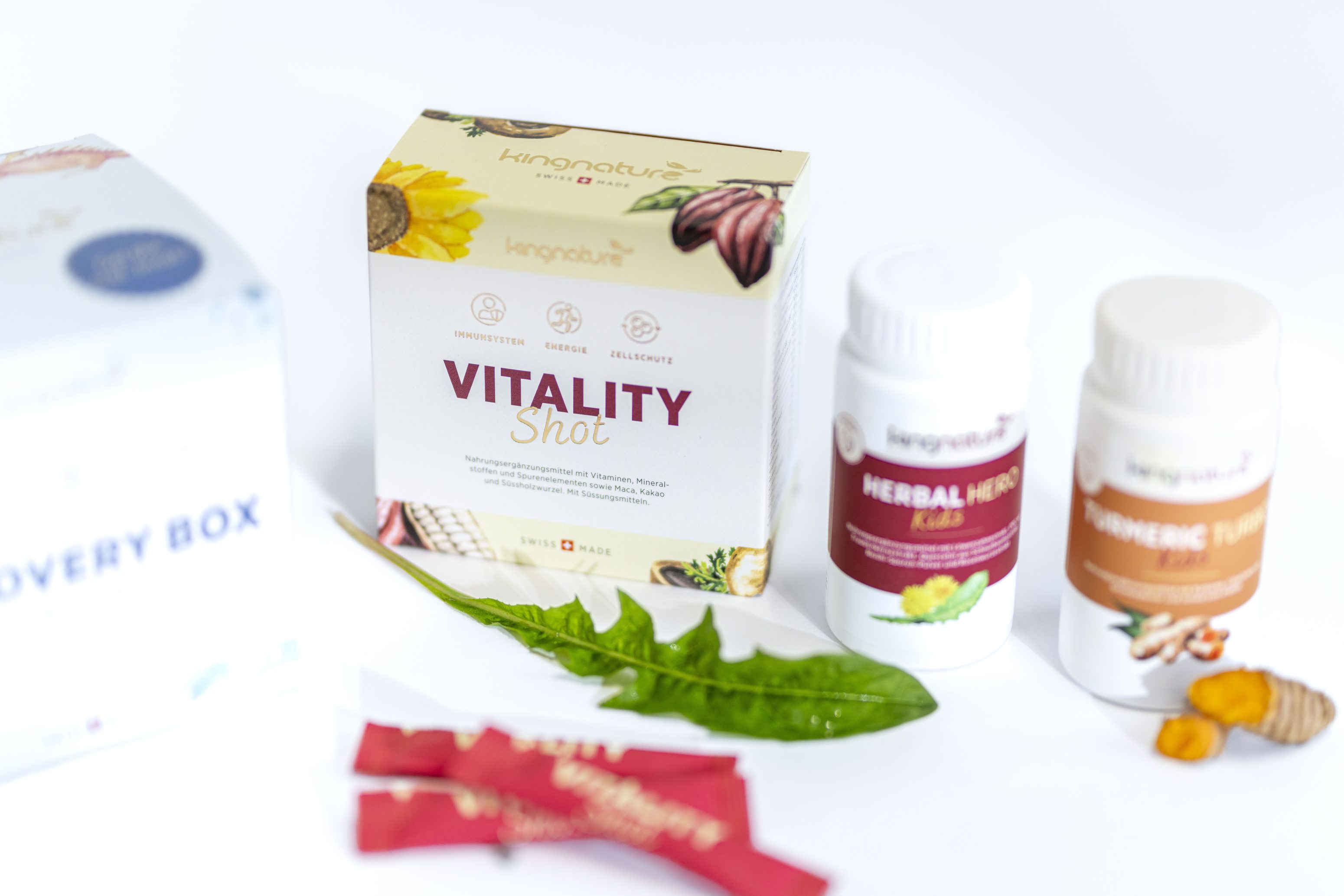 Eine Schachtel mit der Aufschrift Vitality Shot, zwei Fläschchen mit Nahrungsergänzungsmitteln, zwei rote Tütchen und grüne Blätter sind auf einer weißen Fläche mit weißem Hintergrund angeordnet.