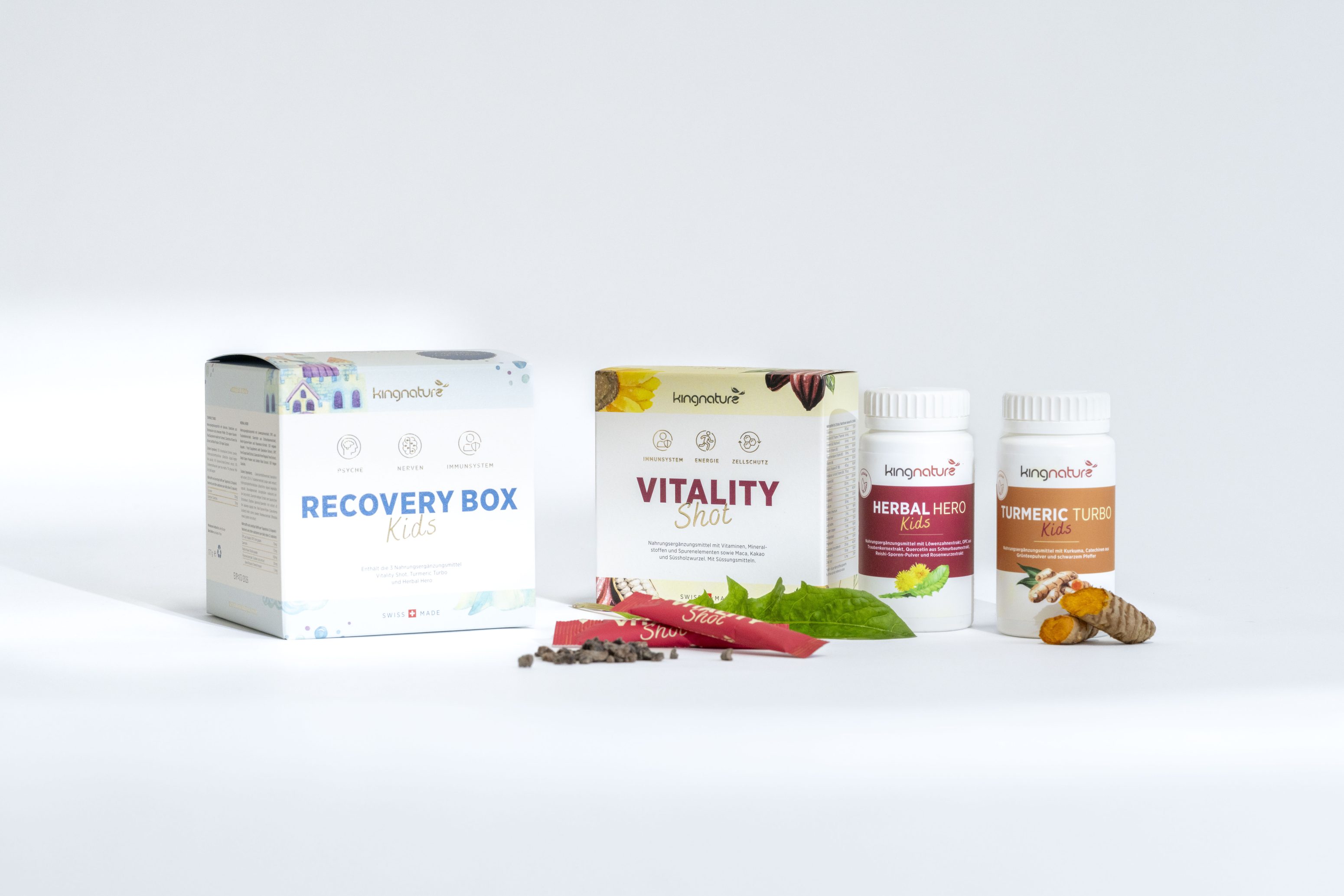 Es werden vier Ergänzungsprodukte vorgestellt: Recovery Box Kids, Vitality Shot, Herbal Hero Defense und Turmeric Tussis. Einige natürliche Inhaltsstoffe wie Kurkumawurzel und -blätter sind vor den Produkten auf einem weißen Hintergrund angeordnet.