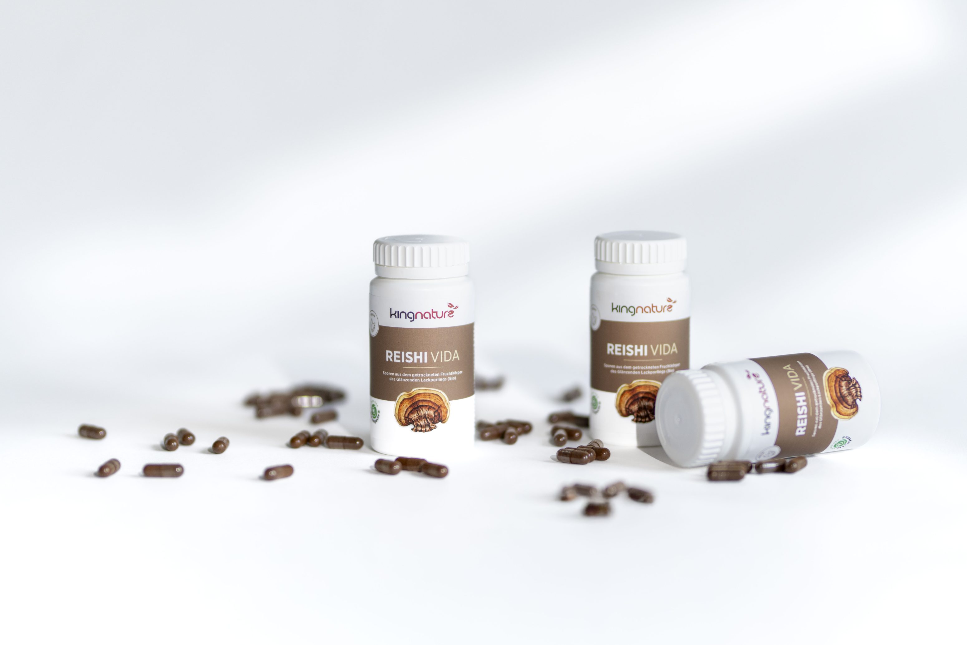 Drei Flaschen mit Kingnature Reishi Vida Nahrungsergänzungsmitteln sind auf einer weißen Fläche ausgestellt, umgeben von verstreuten dunklen Kapseln. Zwei Flaschen stehen aufrecht, und eine liegt auf der Seite.