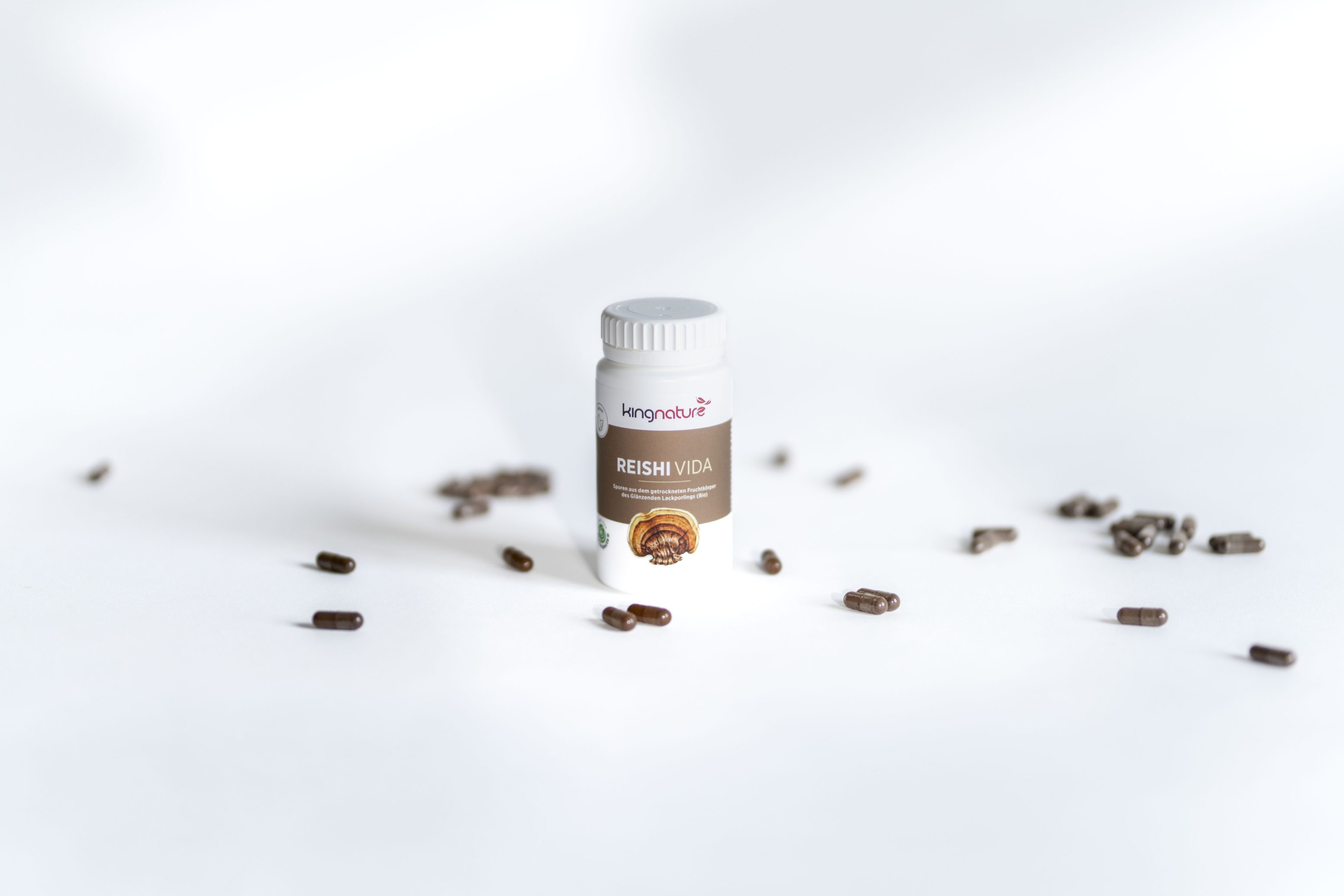 Eine kleine weiße Flasche mit der Aufschrift Reishi Vida steht aufrecht auf einer weißen Fläche, umgeben von verstreuten braunen Kapseln. Die Flasche hat eine weiße Kappe und ein Etikett mit der Abbildung eines Pilzes.
