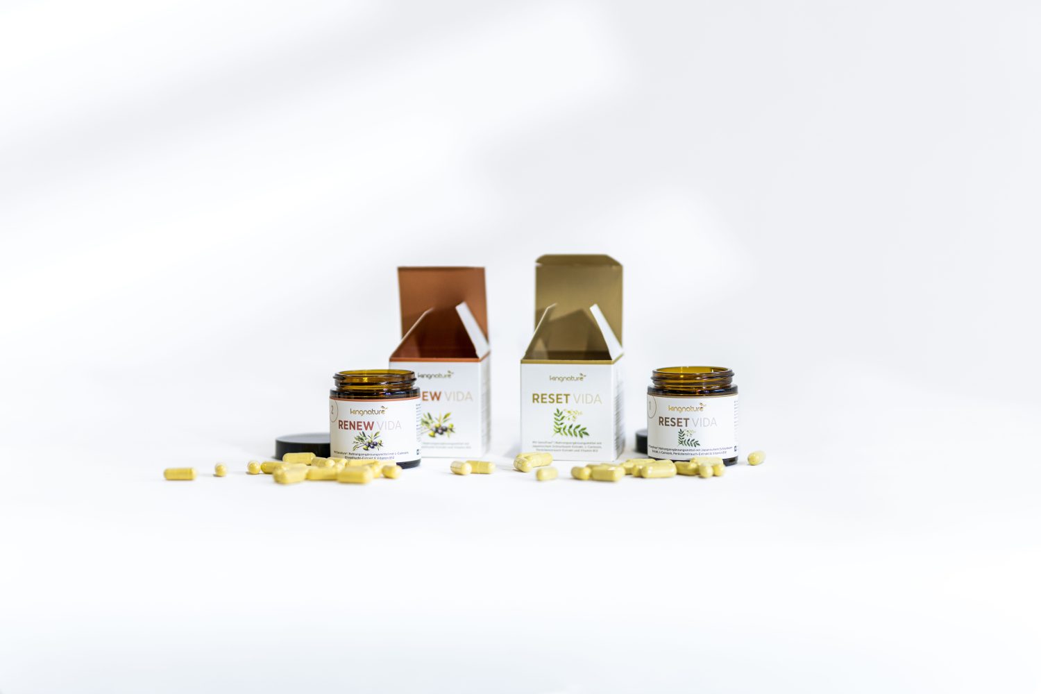 Zwei braune Gläser mit den Aufschriften RENEW VIDA und RESET VIDA stehen vor ihren geöffneten Schachteln, um die herum gelbe Kapseln auf weißem Grund verstreut sind.
