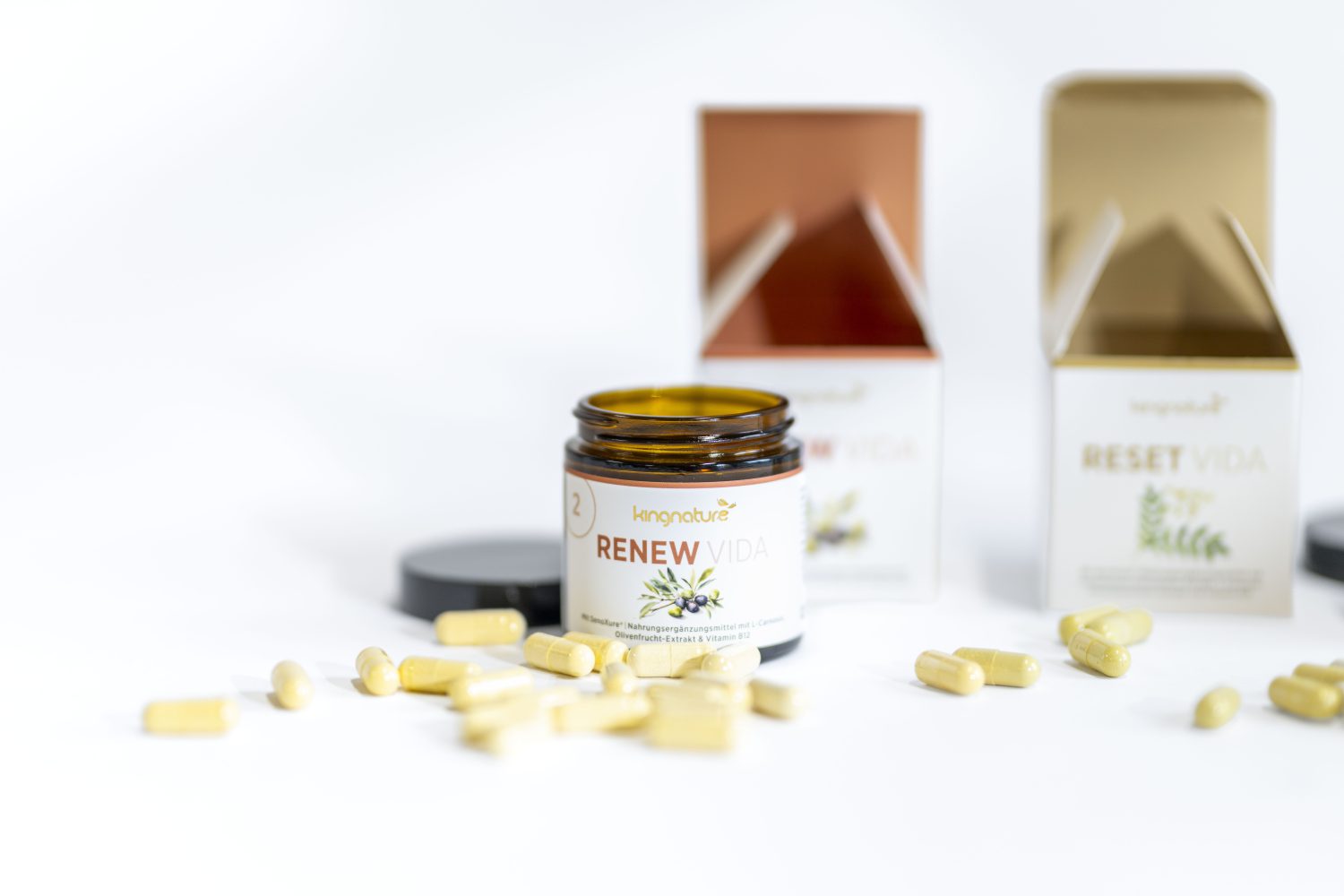 Ein Glas mit der Aufschrift RENEW VIDA, vor dem gelbe Kapseln verschüttet sind, daneben zwei geöffnete Schachteln, eine davon mit der Aufschrift RESET VIDA, alles auf weißem Hintergrund.