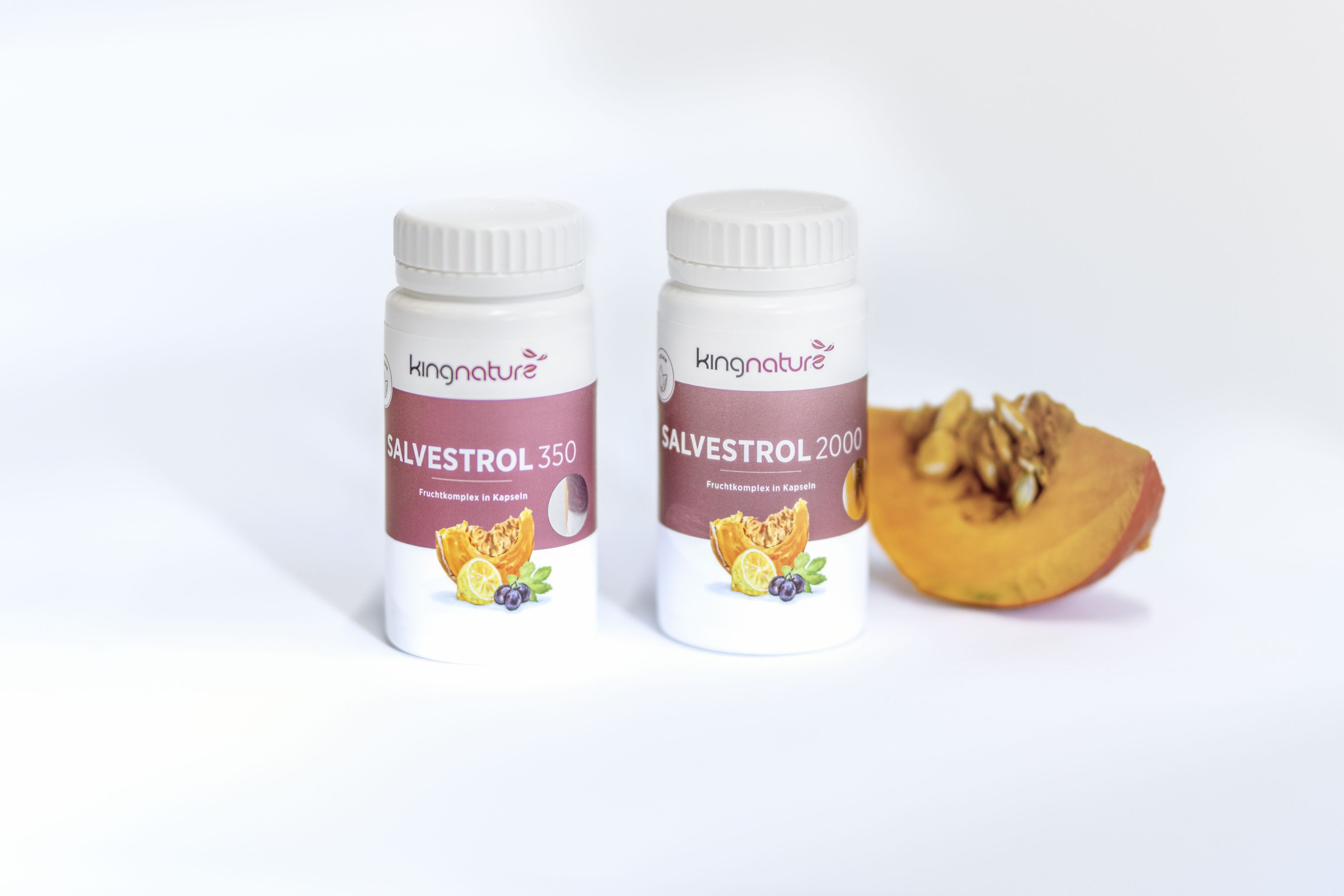 Zwei weiße Flaschen mit Kingnature Salvestrol Nahrungsergänzungsmitteln stehen auf einer weißen Fläche neben einer aufgeschnittenen Orangenfrucht mit einem Kern. Die Flaschenetiketten sind rot und weiß und tragen die Aufschrift Salvestrol 350 und Salvestrol 2000.