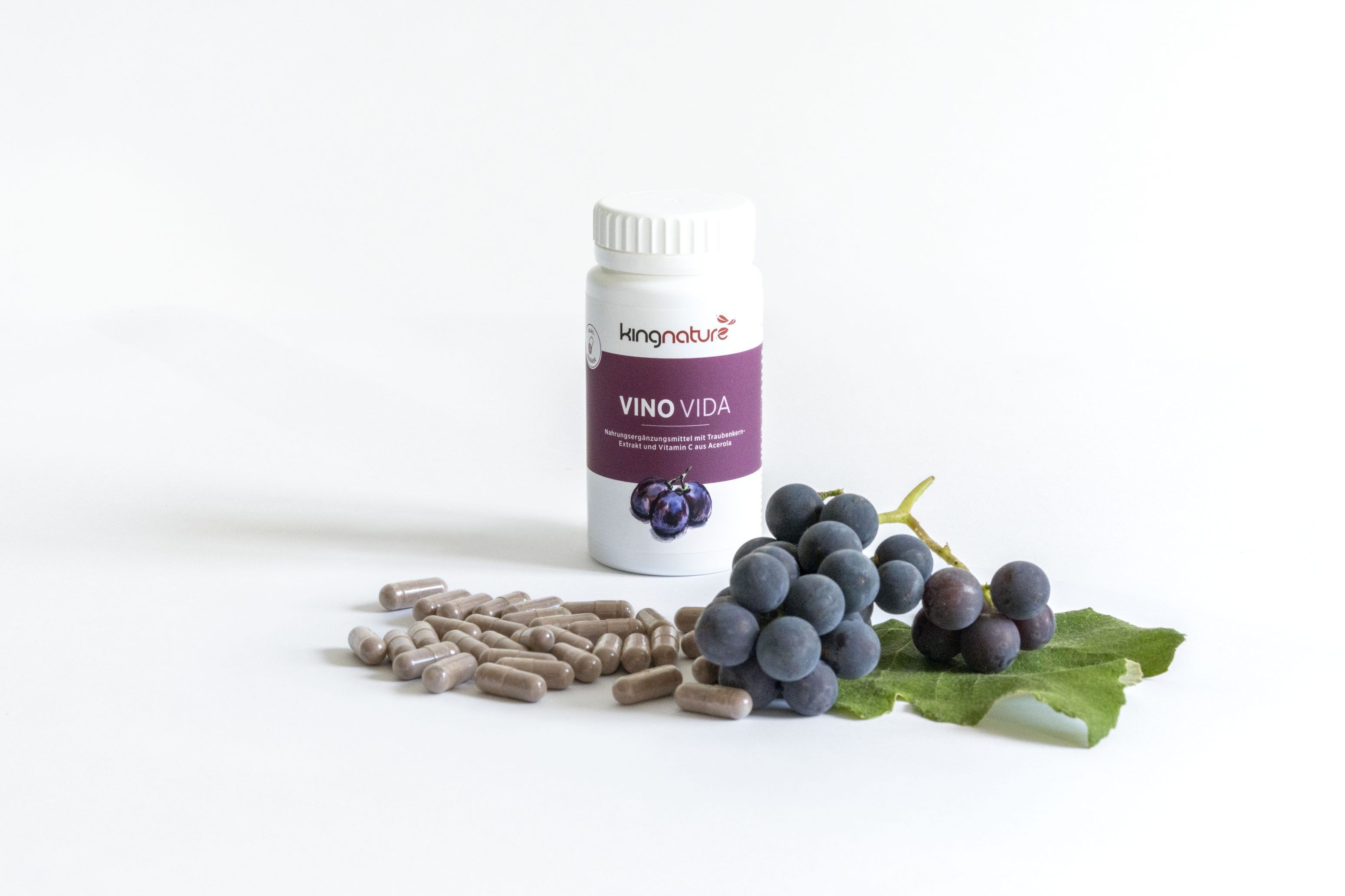 Eine Flasche mit der Aufschrift VINO VIDA steht hinter einem Stapel Kapseln und einem Strauß frischer violetter Trauben mit grünen Blättern auf weißem Hintergrund.