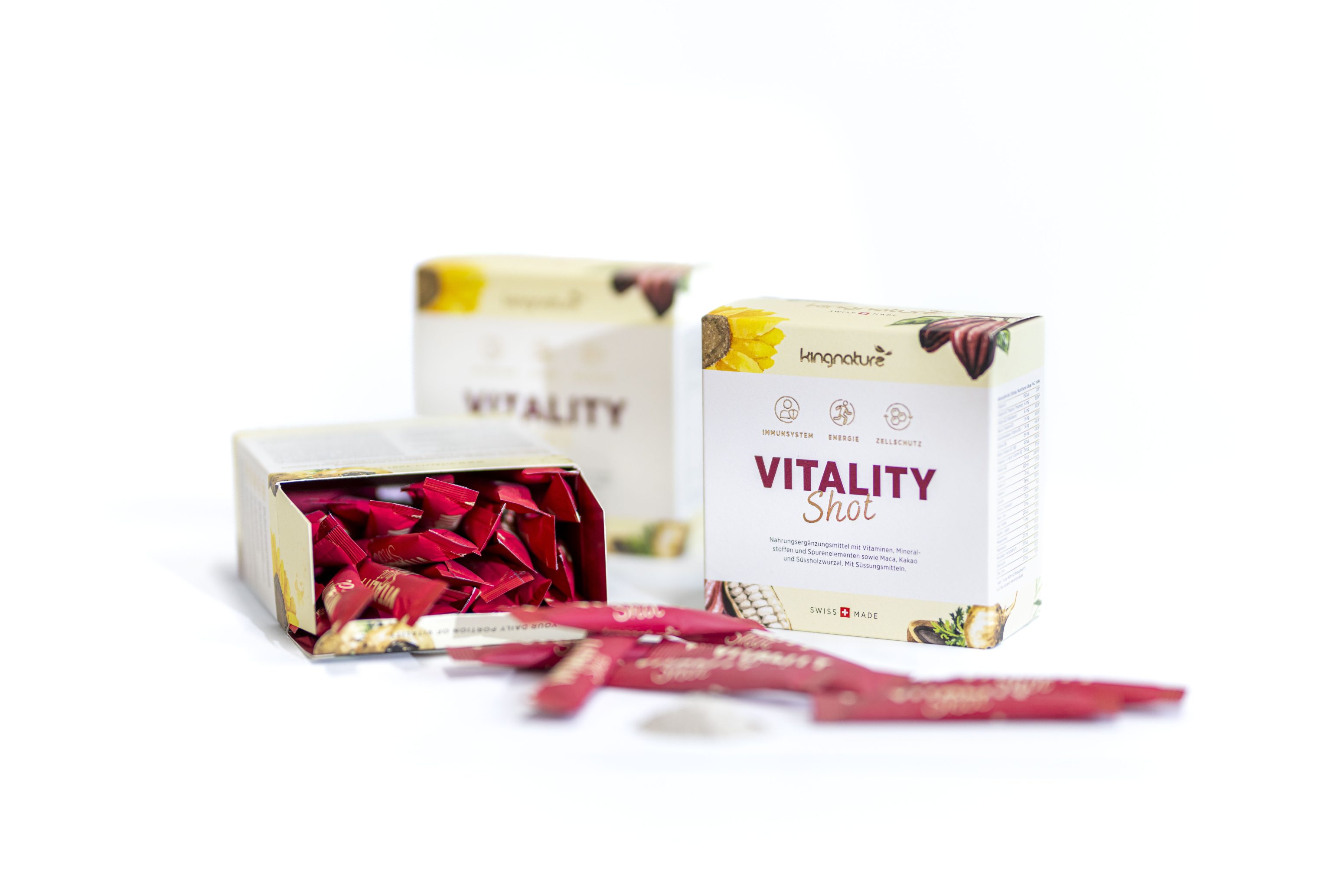Es werden drei Schachteln Kingnature Vitality Shot gezeigt. Eine Schachtel ist geöffnet und enthüllt rote Beutel, während im Vordergrund ebenfalls Beutel verstreut sind. Die Schachteln haben ein weißes Design mit bunten Fruchtabbildungen.