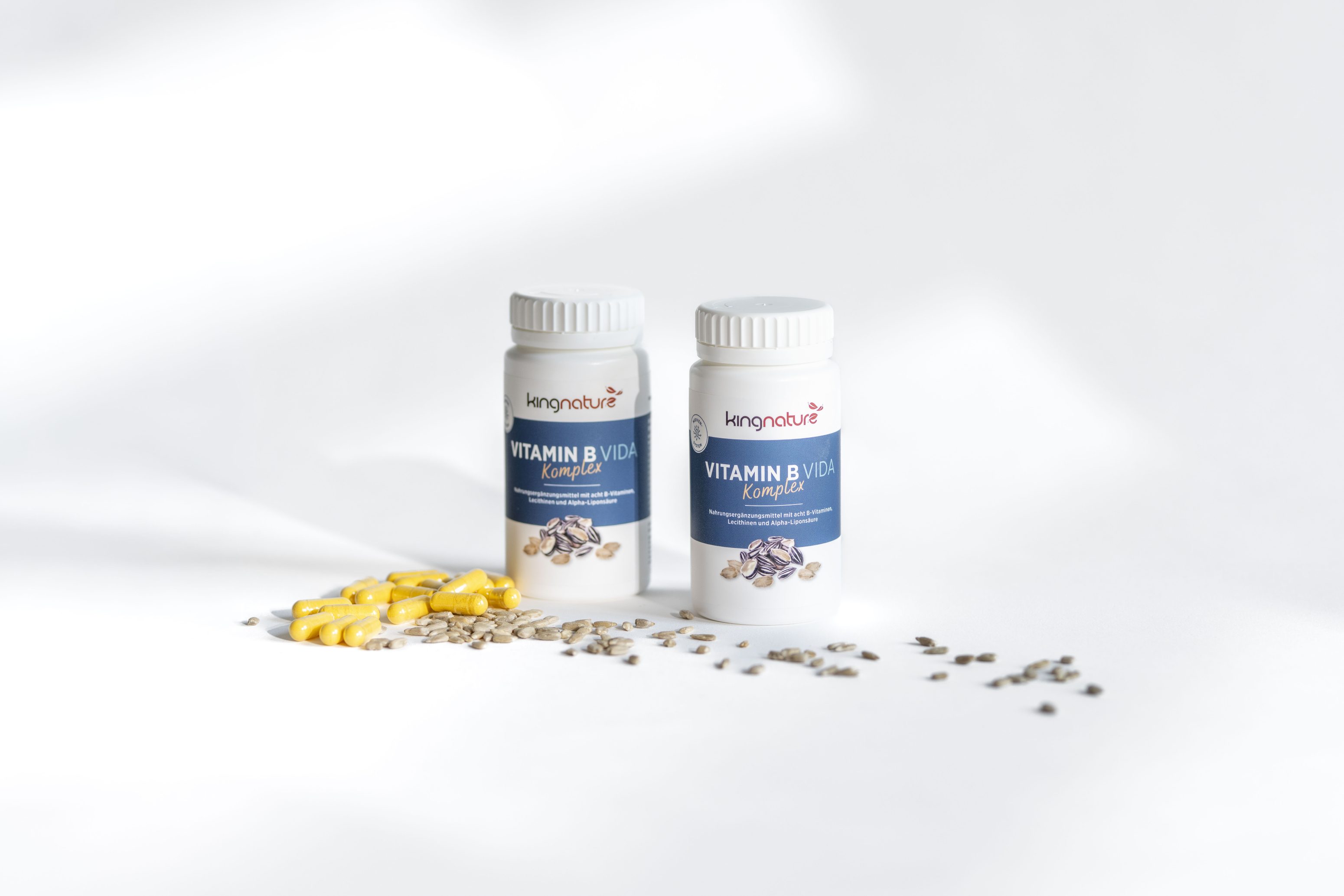 Zwei Flaschen mit der Aufschrift VITAMIN B KOMP stehen auf einer weißen Fläche, vor denen gelbe Kapseln und Samen verstreut sind. Auf den Etiketten sind Nüsse und Samen abgebildet. Das Bild ist hell und hat einen minimalen Hintergrund.