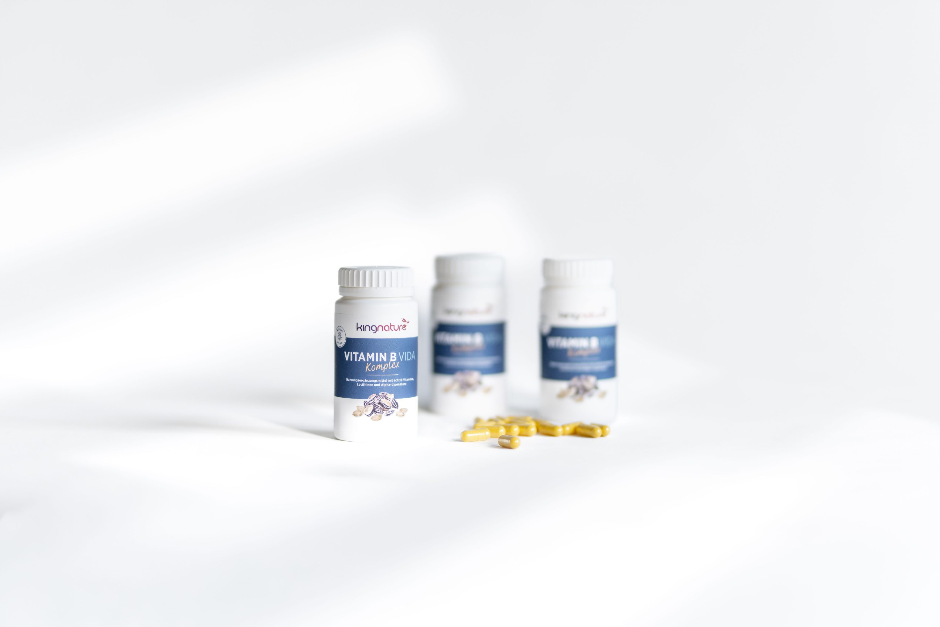 Drei weiße Flaschen mit Vitamin-D3-Präparaten mit blauen Etiketten sind auf einer weißen Fläche aufgestellt. Zwei gelbe Softgel-Kapseln liegen vor den Flaschen. Der Hintergrund ist hell und minimalistisch.