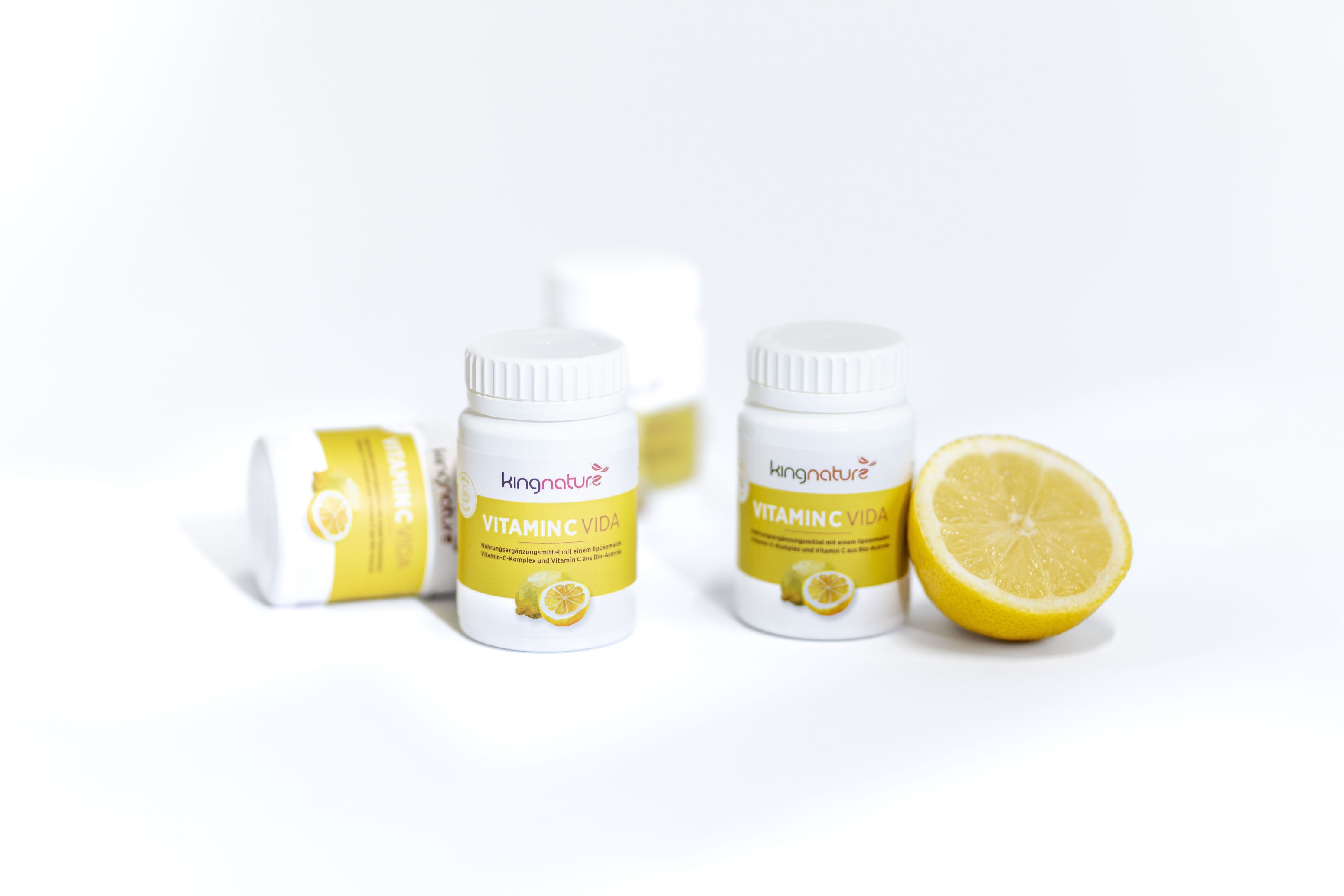 Zwei weiße Flaschen mit der Aufschrift Vitamin C Vida und gelben Etiketten und Zitronenbildern stehen neben einer halbierten Zitrone; zwei weitere Flaschen liegen unscharf im Hintergrund auf einer weißen Fläche.