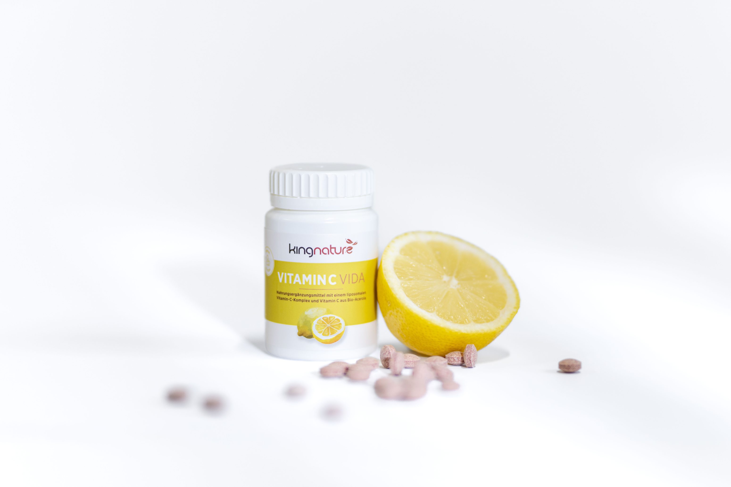 Eine Flasche mit Vitamin-C-Präparaten von Living Nature steht neben einer halbierten Zitrone, auf der mehrere runde rosa Tabletten auf einer weißen Fläche verstreut sind.