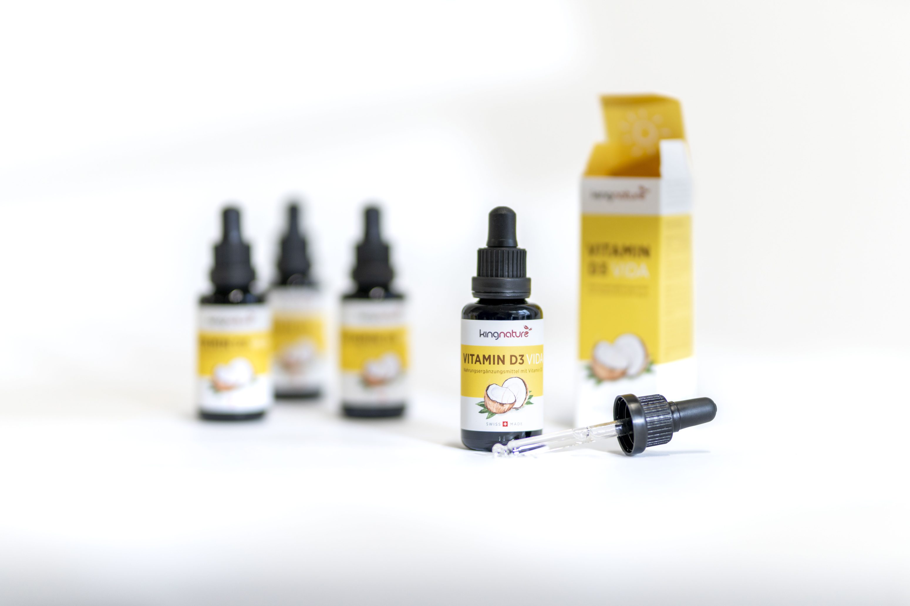 Drei unscharfe Flaschen und eine scharfe Flasche mit Vitamin-D3-Tropfen, mit einem Tropfer und einer offenen gelb-weißen Schachtel mit der Aufschrift Vitamin D3 Vega auf einer weißen Fläche.