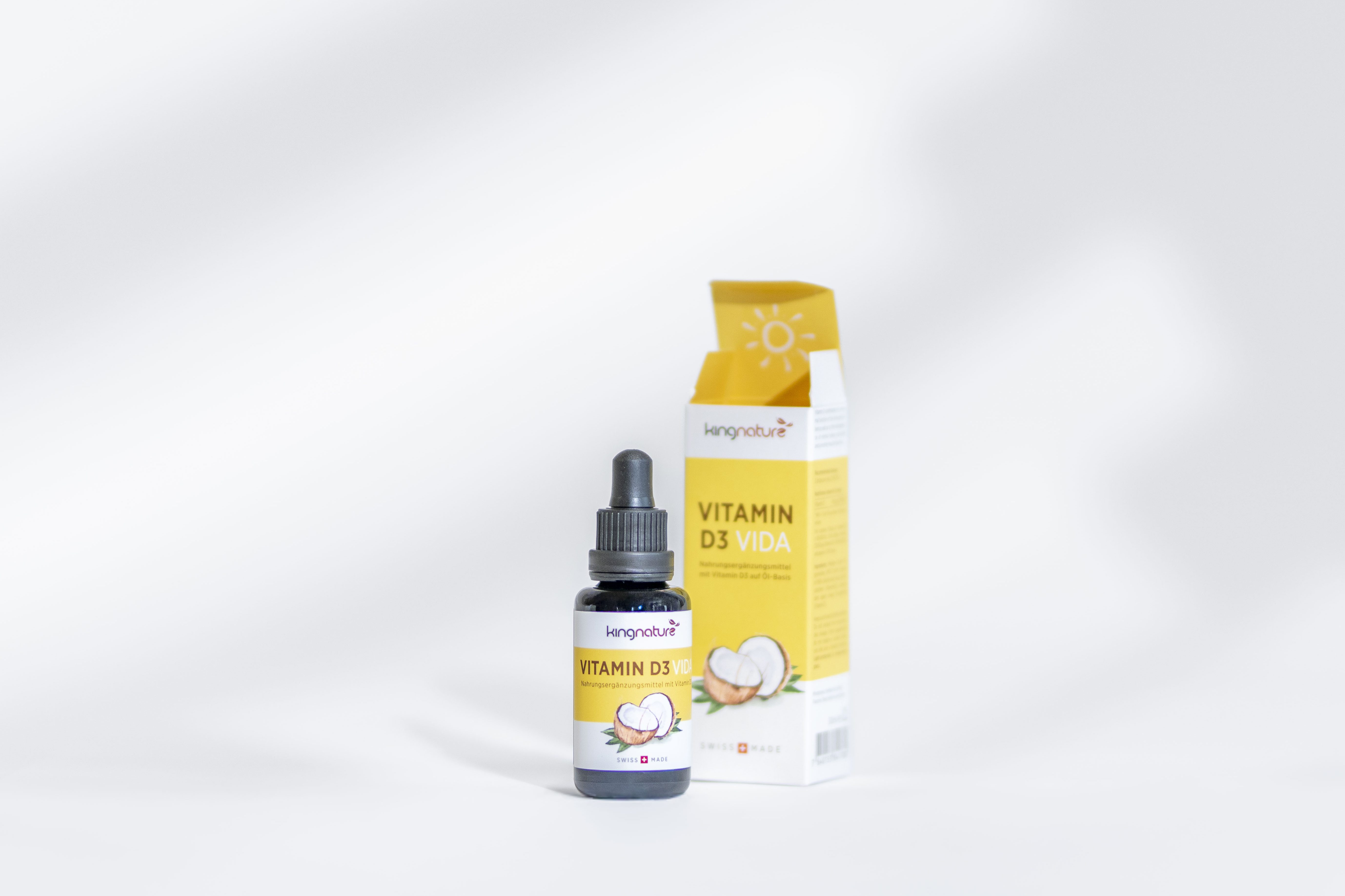 Ein kleines Fläschchen mit einem Tropfer für Vitamin D3 steht vor seiner gelb-weißen Verpackung, die beide mit einer Zitronengrafik versehen sind, auf einer weißen Fläche mit weichen Schatten.