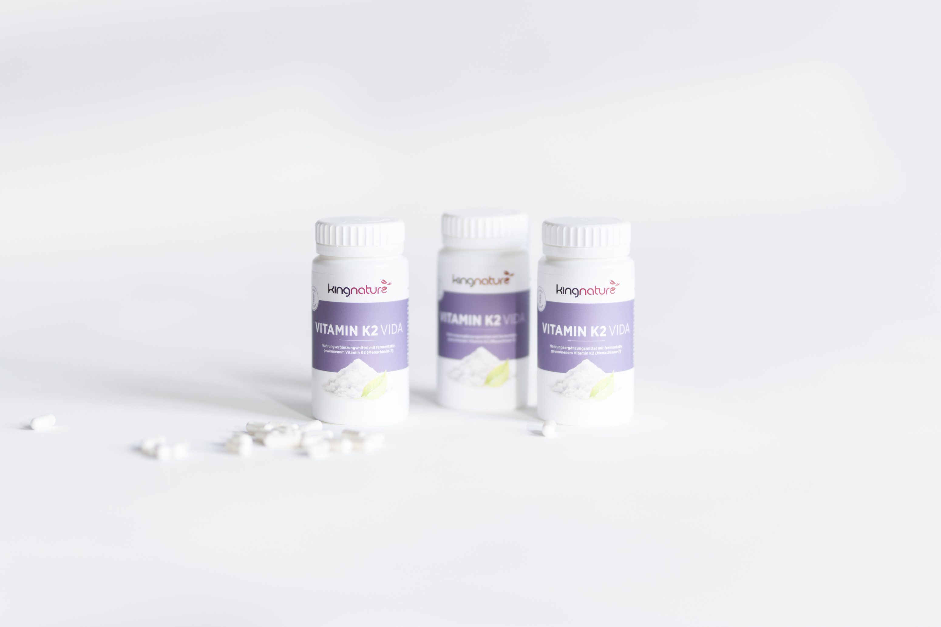Drei weiße Flaschen mit der Aufschrift Vitamin K2 | D3 mit lila Etiketten und weißen Verschlüssen stehen aufrecht auf einer weißen Fläche. Ein paar Pillen sind im Vordergrund verstreut. Der Hintergrund ist hell und minimalistisch.