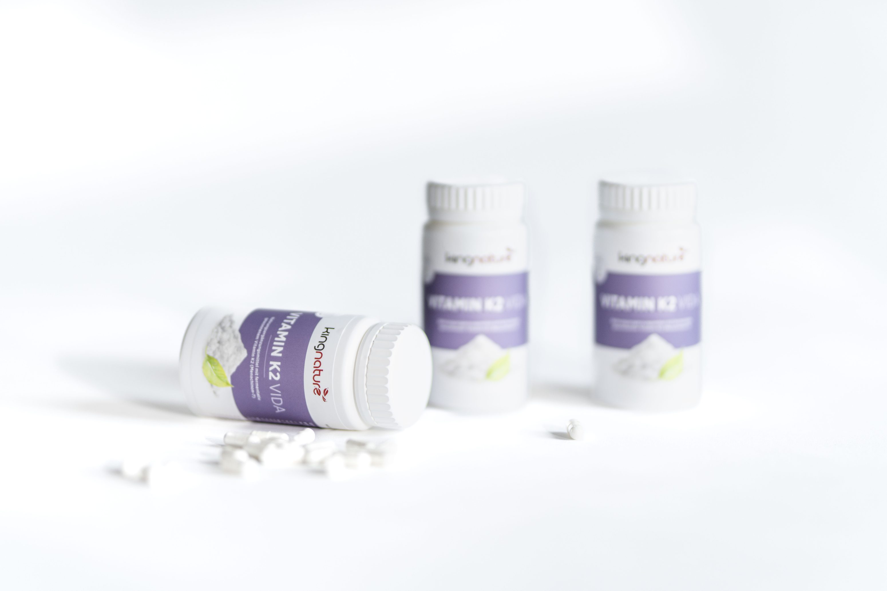 Drei Flaschen mit Vitamin-K2-Ergänzungspräparaten, von denen eine auf der Seite liegt und mehrere weiße Tabletten enthält, und zwei aufrecht im Hintergrund auf einer weißen Fläche stehen.