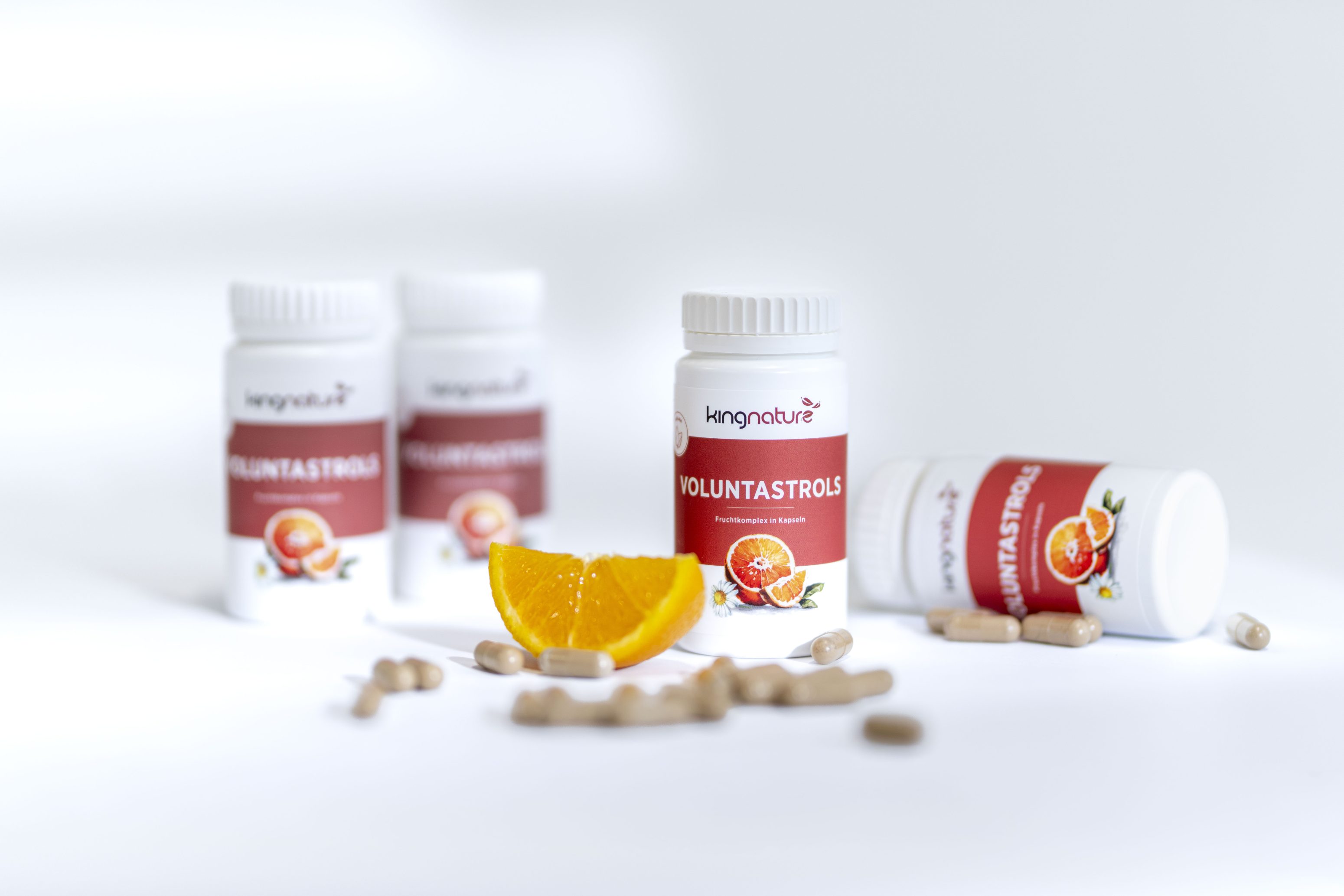 Vier Flaschen mit Voluntastrols-Nahrungsergänzungsmitteln sind auf einer weißen Fläche mit einem aufgeschnittenen Orangenkeil und verstreuten Kapseln im Vordergrund angeordnet. Auf dem Etikett der Flaschen ist eine Orange abgebildet.