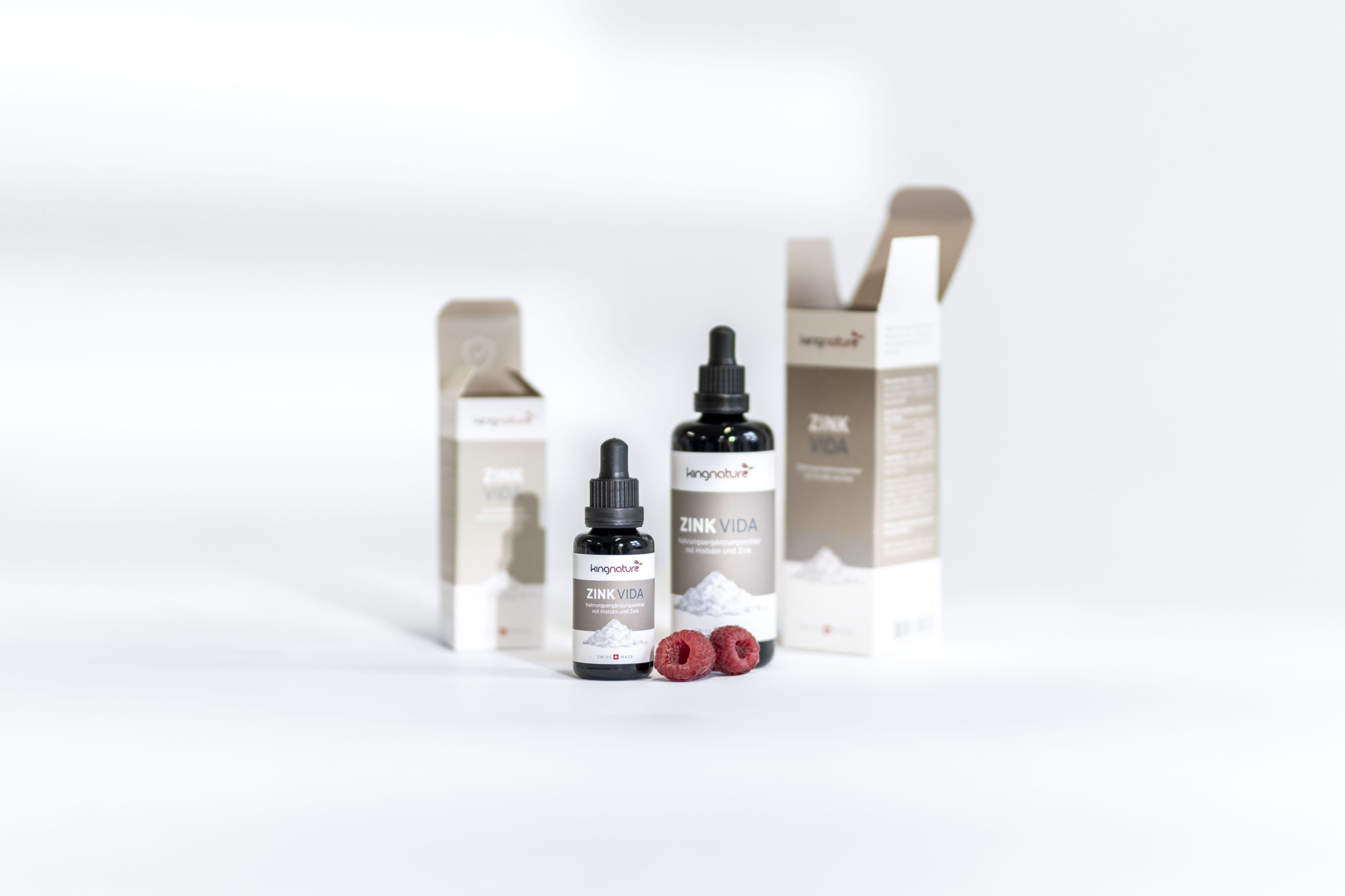 Im Mittelpunkt stehen zwei Tropfflaschen mit der Aufschrift ZINK VIDA, von denen eine offen und eine geschlossen ist. Dahinter befinden sich zwei weiße und braune Produktschachteln und einige Himbeeren auf einer weißen Fläche.