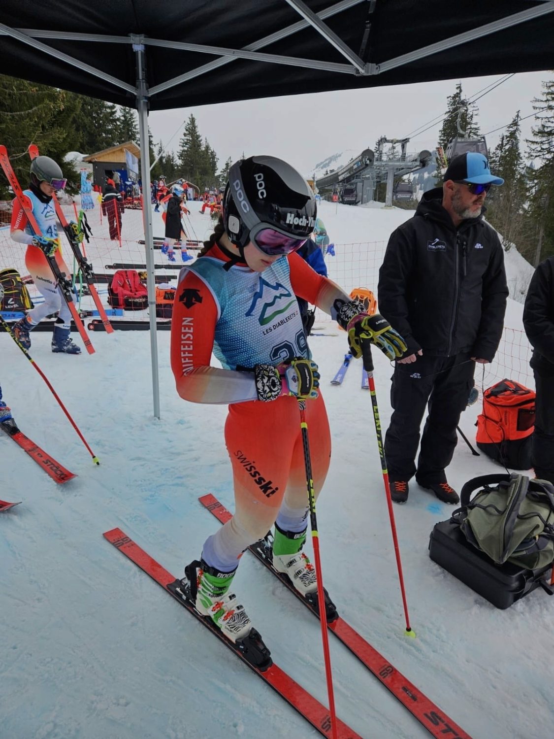 Allegra Frei, eine Skirennfahrerin in einem farbenfrohen Anzug und mit Helm, steht auf Skiern in einem verschneiten Startbereich und justiert ihre Handschuhe und Stöcke, während andere Teilnehmer und ein Offizieller in Winterkleidung in der Nähe unter einem Zelt stehen.