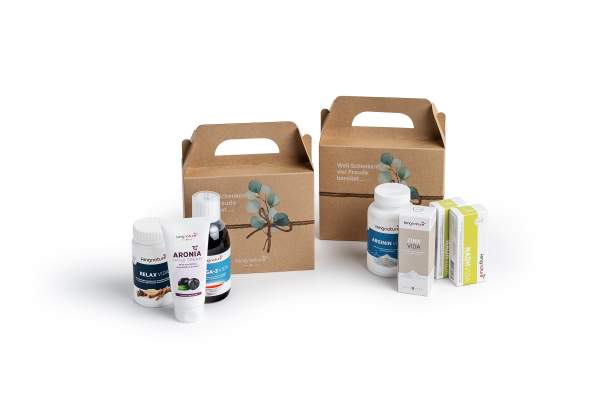 Zwei Geschenk-Boxen aus Karton mit Eukalyptus-Design sind von verschiedenen Flaschen und Schachteln mit Nahrungsergänzungsmitteln umgeben, darunter Vitamine, ein Nasenspray und ein Aroniabeerenprodukt, alles vor einem weißen Hintergrund.