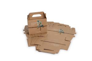 Eine kleine braune Geschenkbox aus Karton mit Henkel und aufgedrucktem Pflanzenmotiv steht aufgebaut vor mehreren flachen, unmontierten Kraftkartons.