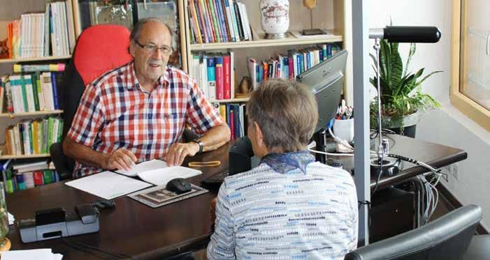 Ein älterer Mann im karierten Hemd sitzt an einem Schreibtisch und unterhält sich mit einer Frau ihm gegenüber im Büro der Naturheilpraxis Landmesser, umgeben von Bücherregalen, Pflanzen und einem Computer.