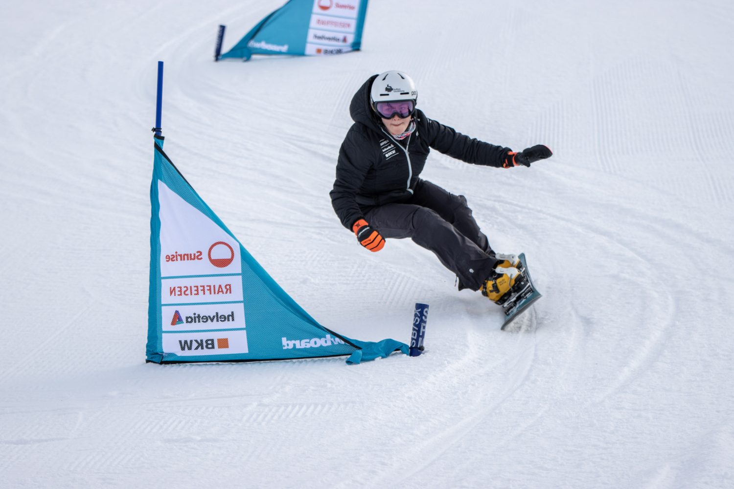 Ein Snowboarder in schwarzer Ausrüstung und weißem Helm fährt scharf um ein blaues Slalomtor auf einer verschneiten Strecke und wirbelt Schnee auf, während er sich in die Kurve legt – eine Erinnerung an den dynamischen Rennstil von Ricarda Hauser.