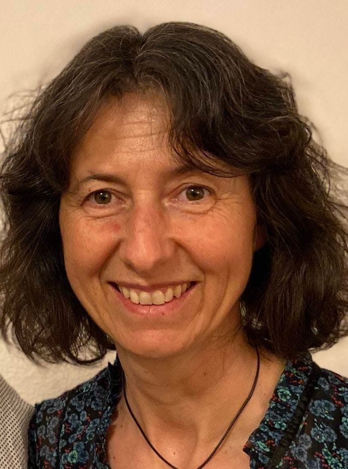 Eine Frau mit welligem braunem Haar lächelt in die Kamera. Gesundheitscoach Sonja Crepaldi trägt ein dunkles, gemustertes Oberteil und eine schwarze Halskette und steht vor einem schlichten, hellen Hintergrund.