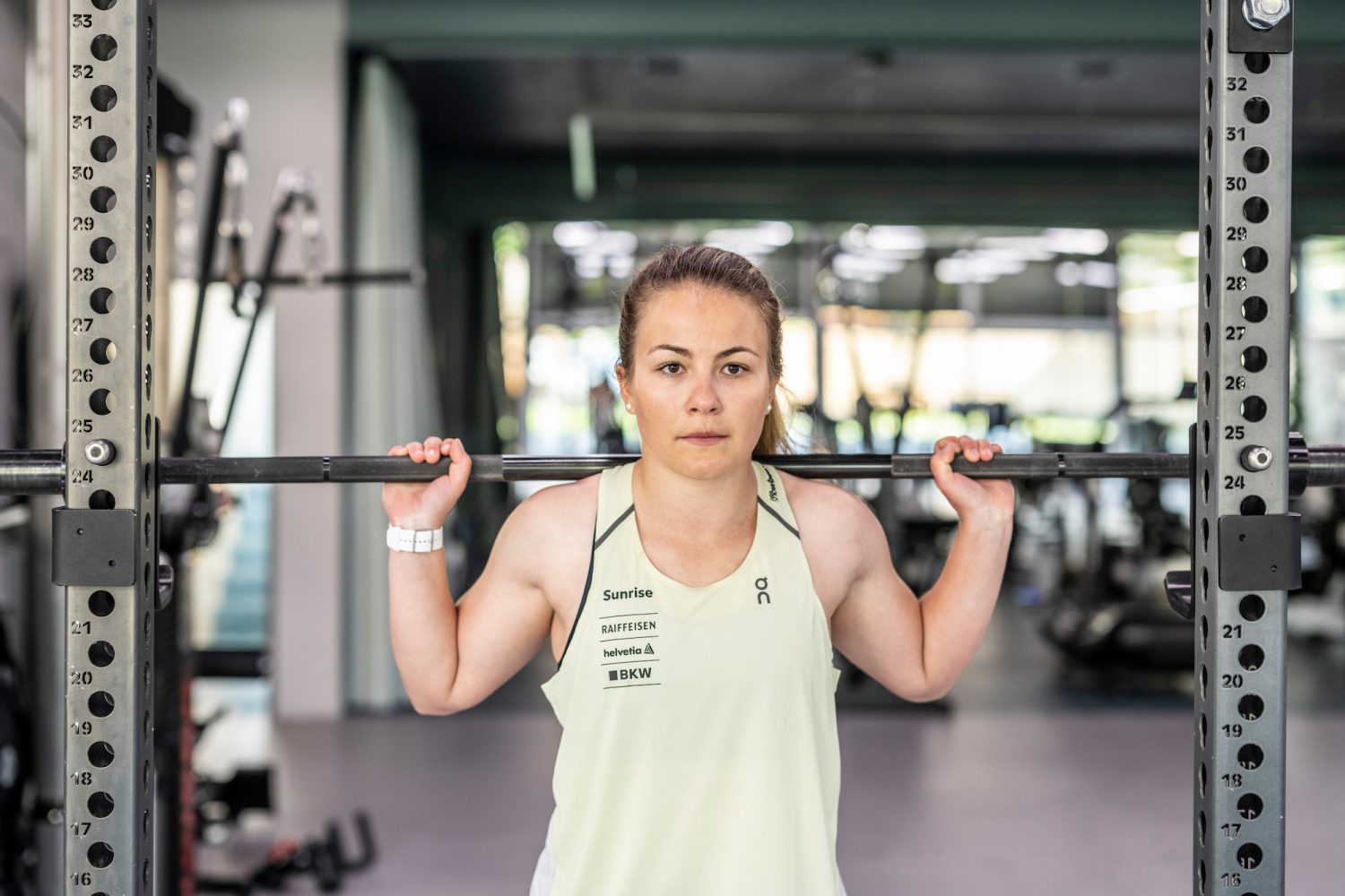 Ricarda Hauser steht mit einer Hantel über den Schultern in einem Fitnessstudio und bereitet sich aufs Gewichtheben vor. Sie trägt ein helles Tanktop und eine Armbanduhr und wirkt konzentriert und entschlossen. Im Hintergrund sind Fitnessgeräte zu sehen.