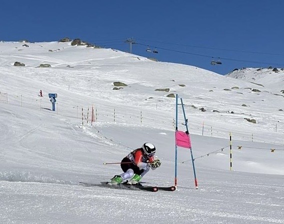 Allegra Frei, eine Skifahrerin in einem rot-schwarzen Anzug und weißem Helm, fährt bergab und biegt in eine Kurve um ein rosa-blaues Slalomtor auf einem verschneiten Berghang unter einem strahlend blauen Himmel ein.