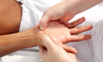 Nahaufnahme einer Person, die einer anderen eine entspannende, therapeutische Handmassage gibt, wobei der Schwerpunkt auf der wohltuenden Berührung liegt. Beide Personen liegen auf einer weißen Fläche, nur ihre Hände und Unterarme sind sichtbar.
