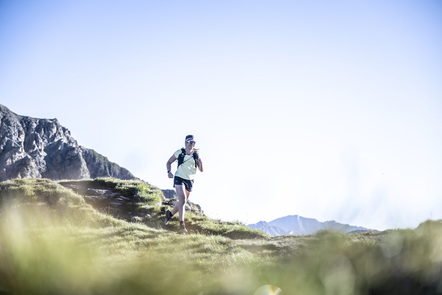 Ricarda Hauser läuft in Sportkleidung und mit Rucksack auf einem Graspfad in einer Berglandschaft unter einem klaren blauen Himmel, wobei das Sonnenlicht eine helle und lebendige Szenerie schafft.