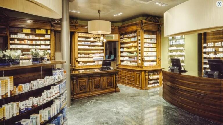 Die Bellevue Apotheke ist eine moderne Apotheke mit eleganten Holzregalen und Theken, gut beleuchtet und mit Gesundheitsprodukten bestückt. Zwei Computerstationen und Topfpflanzen sorgen für eine warme, einladende Atmosphäre.