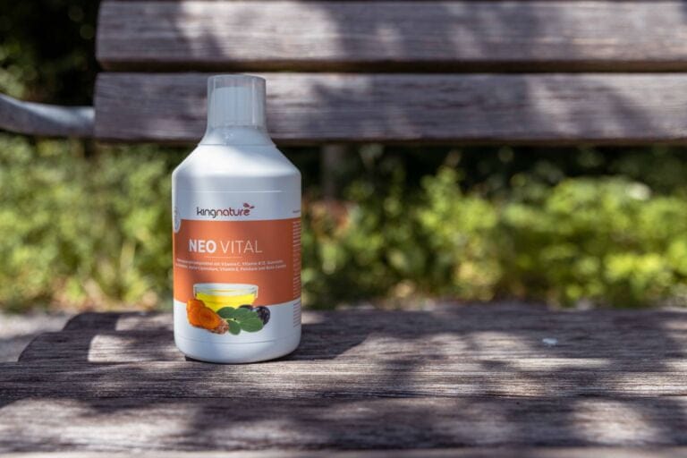 Eine Flasche Kingnature Neovital steht auf einer Holzbank im Freien, im Hintergrund sind Sonnenlicht und Grünpflanzen zu sehen. Das orange-weiße Etikett zeigt Abbildungen von Obst, Gemüse, Alpha-Liponsäure und Vitamin B12.