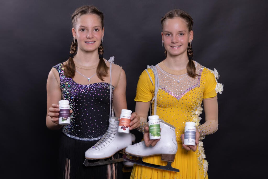 Selina Arnold und Alessia Arnold, zwei junge Frauen mit geflochtenen Haaren in Eiskunstlaufkleidern – eine schwarz, eine gelb – halten Paar Schlittschuhe und präsentieren Nahrungsergänzungsmittelflaschen vor einem schlichten schwarzen Hintergrund.