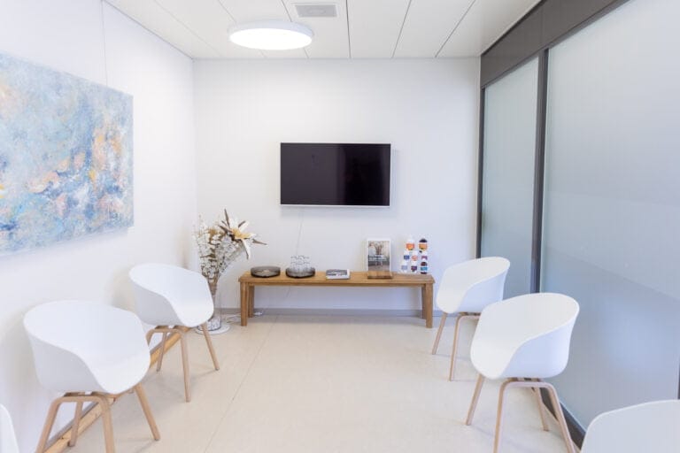 Ein moderner Warteraum im Wise Medicine Gesundheitszentrum verfügt über sechs weiße Stühle, einen an der Wand montierten Fernseher, einen Holztisch mit Zeitschriften und Dekoration, eine Milchglaswand und ein abstraktes Gemälde an der weißen Wand.