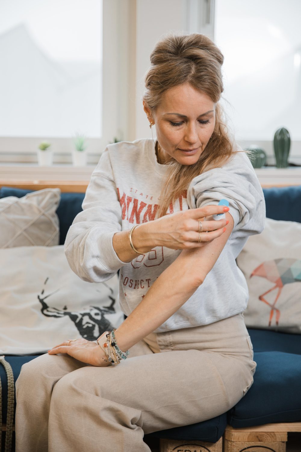 Eine Frau sitzt auf einem Sofa und klebt sich ein rundes Pflaster auf den Oberarm, wobei sie darauf hinunterblickt – sie demonstriert damit einen Wechseljahre-Check für zu Hause. Sie trägt ein lässiges Sweatshirt und eine beige Hose, hinter ihr liegen Zierkissen.
