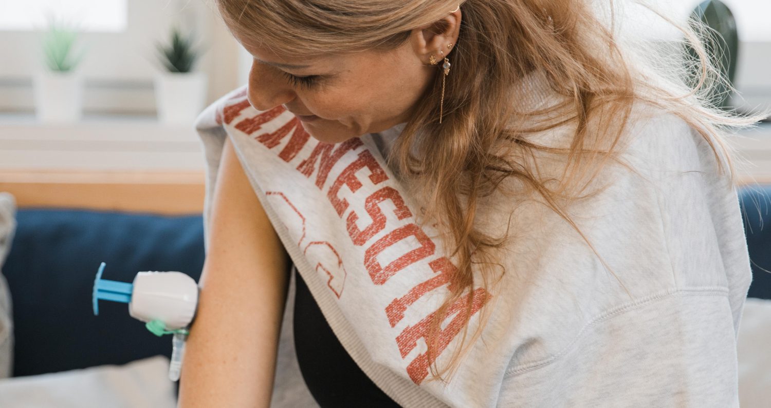 Eine Frau sitzt auf einem Sofa und betrachtet ihren Oberarm, während sie eine Impfung erhält. Sie trägt ein graues Sweatshirt mit hochgekrempelten Ärmeln und hat hellbraunes Haar; möglicherweise bereitet sie sich gerade auf ihren Wechseljahre-Check für zu Hause vor.