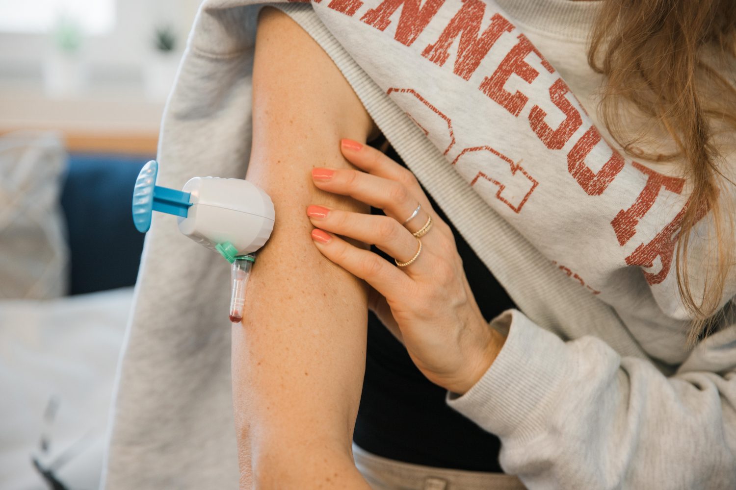Eine Person in einem grauen Sweatshirt nutzt den Herz-Kreislauf-Check zu Hause: Mit dem Gerät entnimmt sie sich Blut aus dem Oberarm, während die andere Hand den Arm stützt, und eine kleine Menge Blut erscheint im Röhrchen.