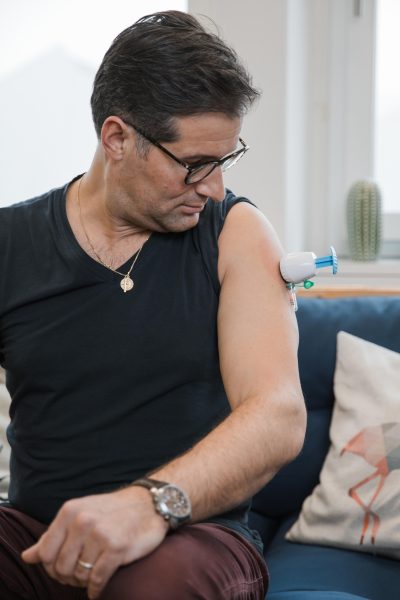 Ein Mann mit Brille und schwarzem Hemd sitzt auf einer blauen Couch und schaut auf ein medizinisches Gerät, das für eine Bleigehaltanalyse an seinem Oberarm befestigt ist. Im Hintergrund sind eine Kaktuspflanze und ein gemustertes Kissen zu sehen.