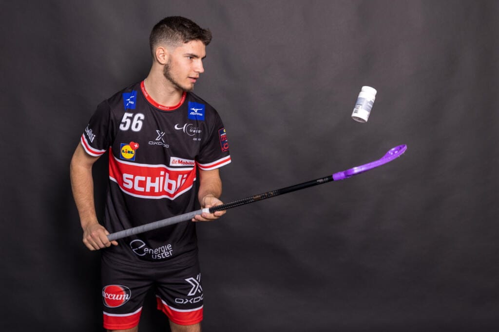 Josia Pfister, ein Unihockeyspieler in schwarz-roter Uniform, wirft mit seinem Schläger vor einem schlichten, dunklen Hintergrund eine weiße Flasche in die Luft.